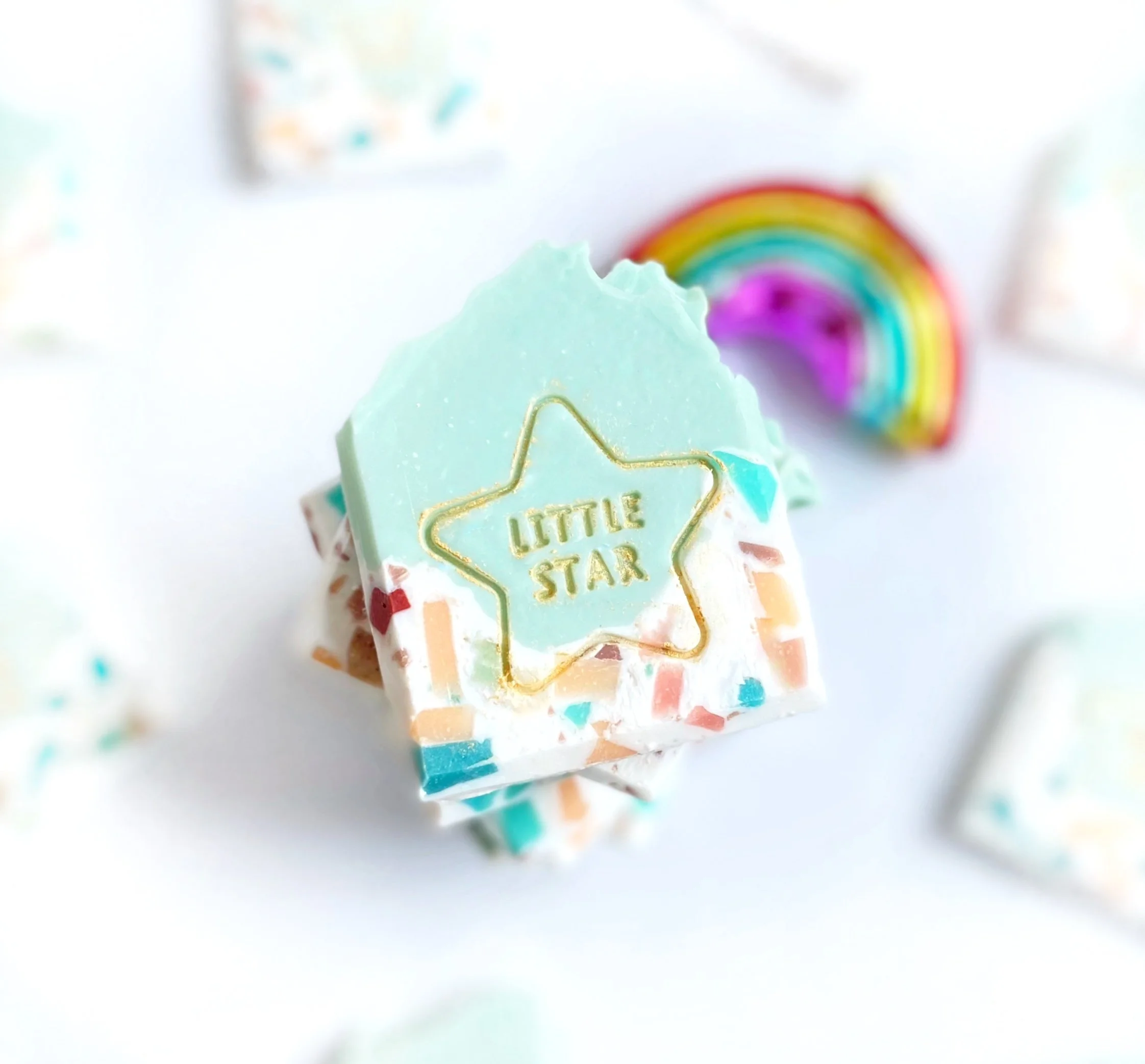 Close-up of a pastel-colored, star-shaped soap with the words "LITTLE STAR" embossed in gold, surrounded by small, multicolored sprinkles, with a blurred rainbow in the background.