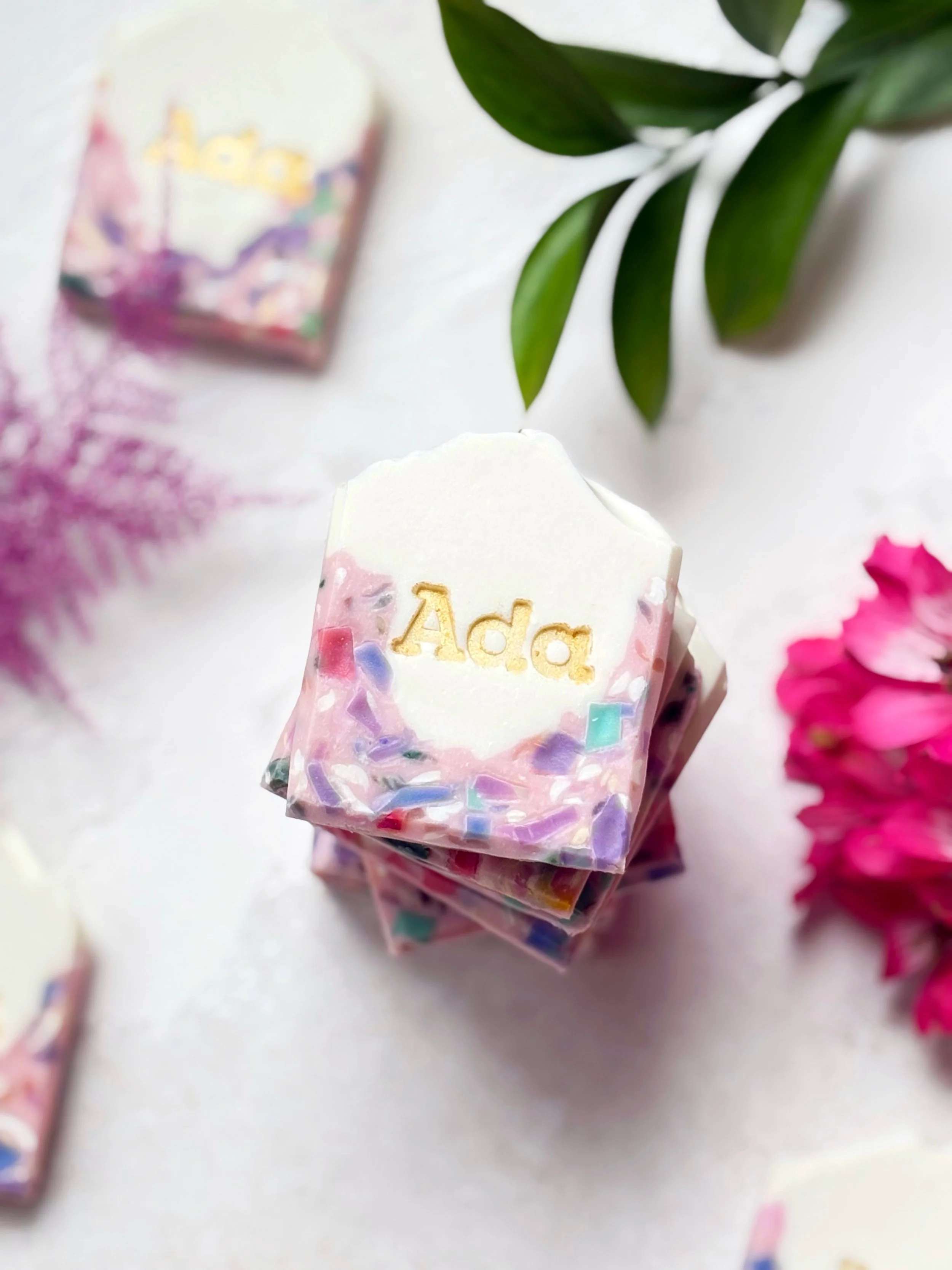 Stack of handcrafted soap bars with the name 'Ada' engraved on the top soap, surrounded by decorative flowers and green leaves.