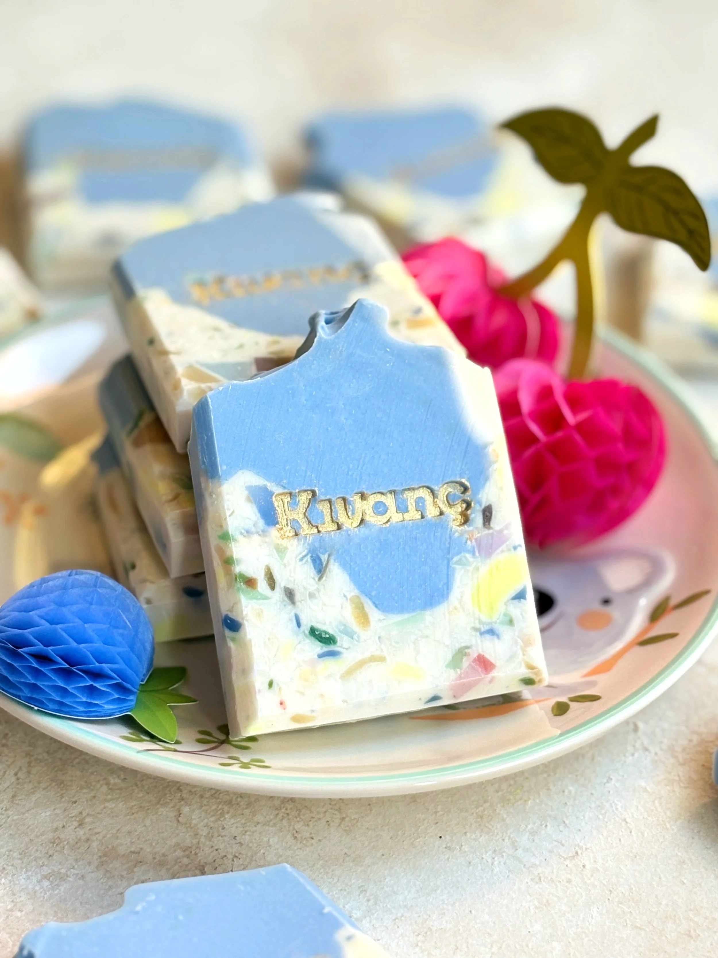 Bar of soap with the word "Kuwang" on a decorative plate, alongside pink paper honeycomb decoration and a blue honeycomb decoration featuring a leaf, with a blurred background.