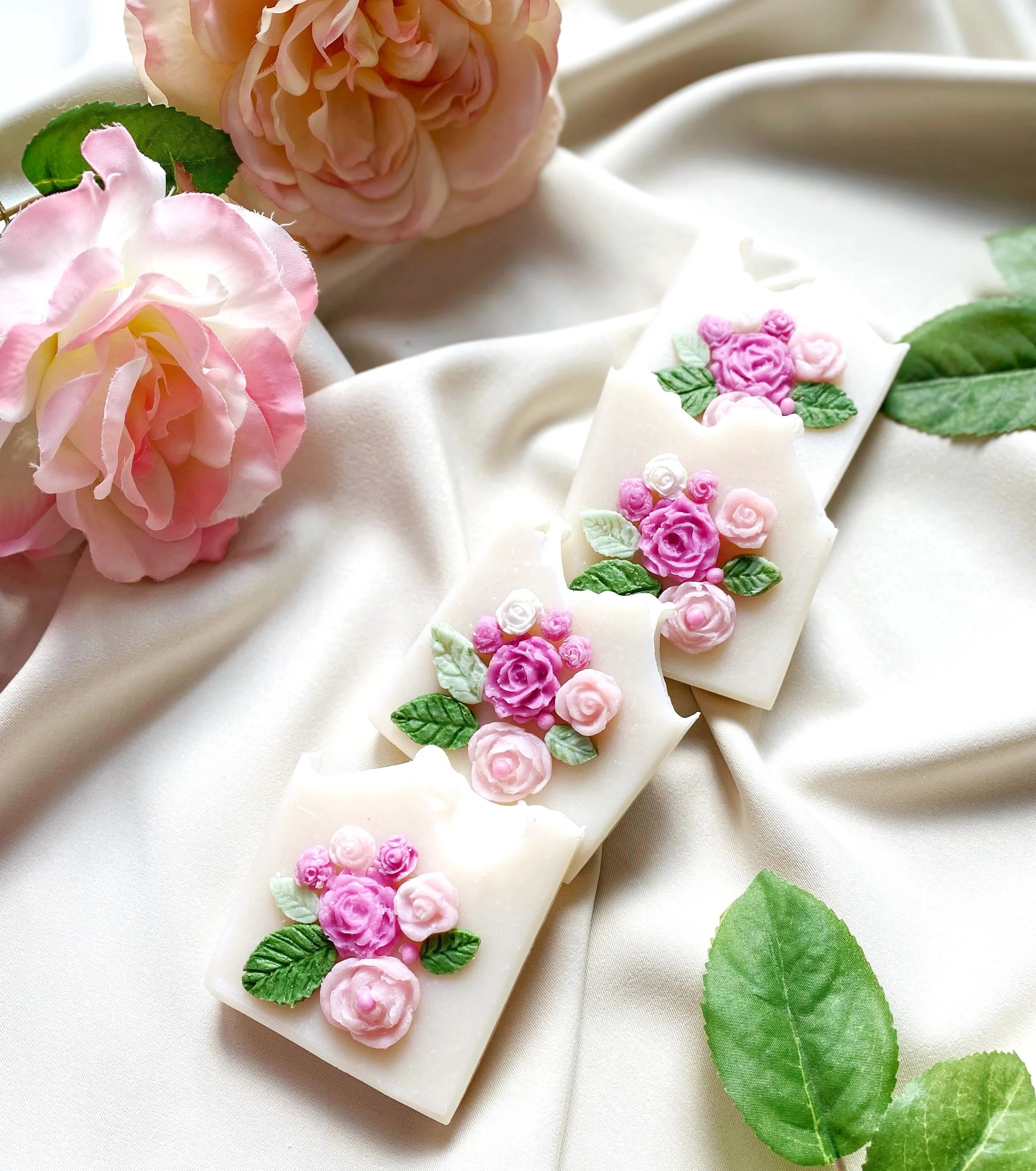 Decorative soap bars with pink and purple rose flower designs, set on white satin fabric with pink roses and green leaves around them.