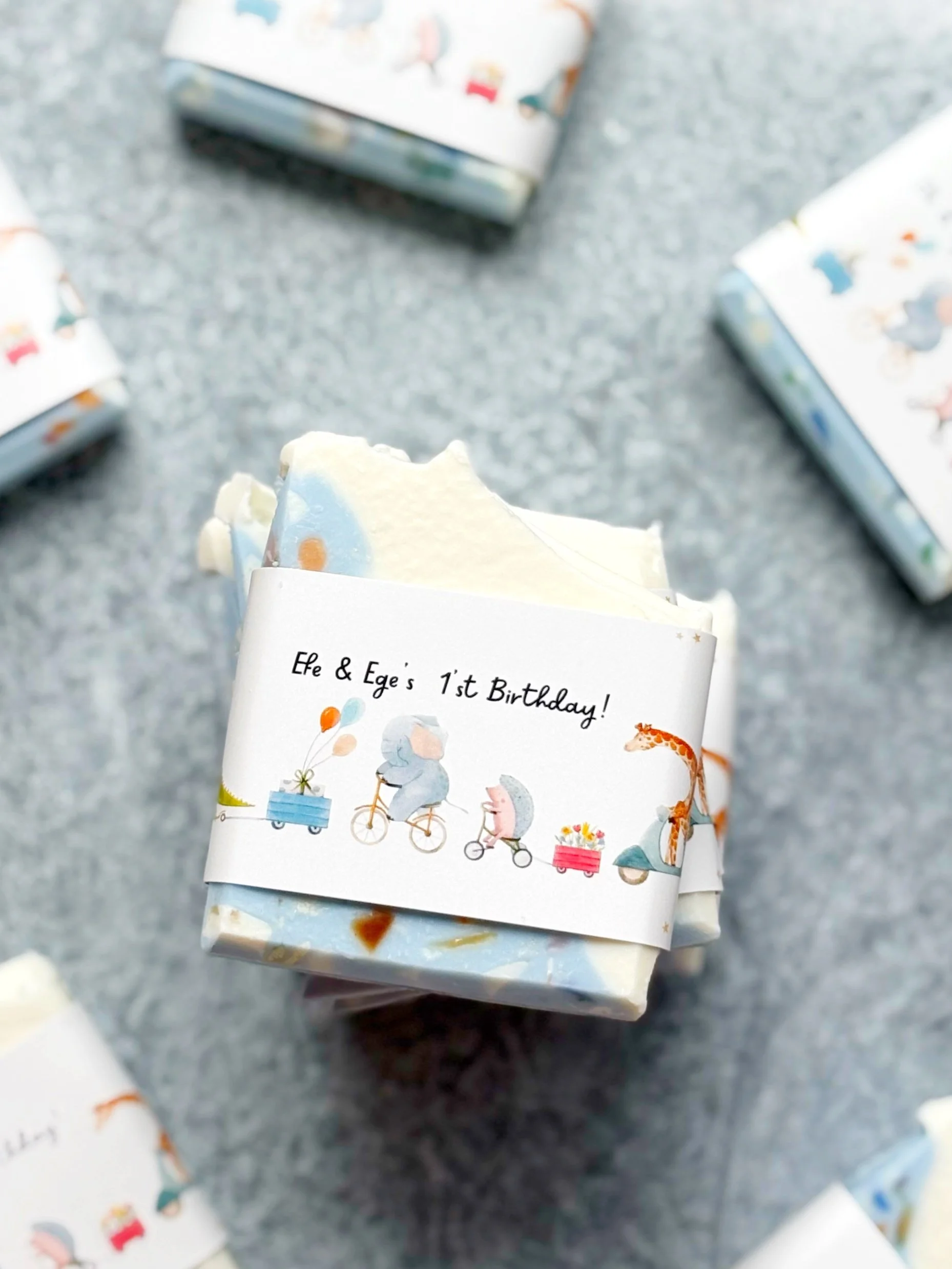 A small gift wrapped in paper with a birthday message for Efe and Ege, featuring illustrations of animals riding bicycles and pulling carts, with a gray textured surface background.
