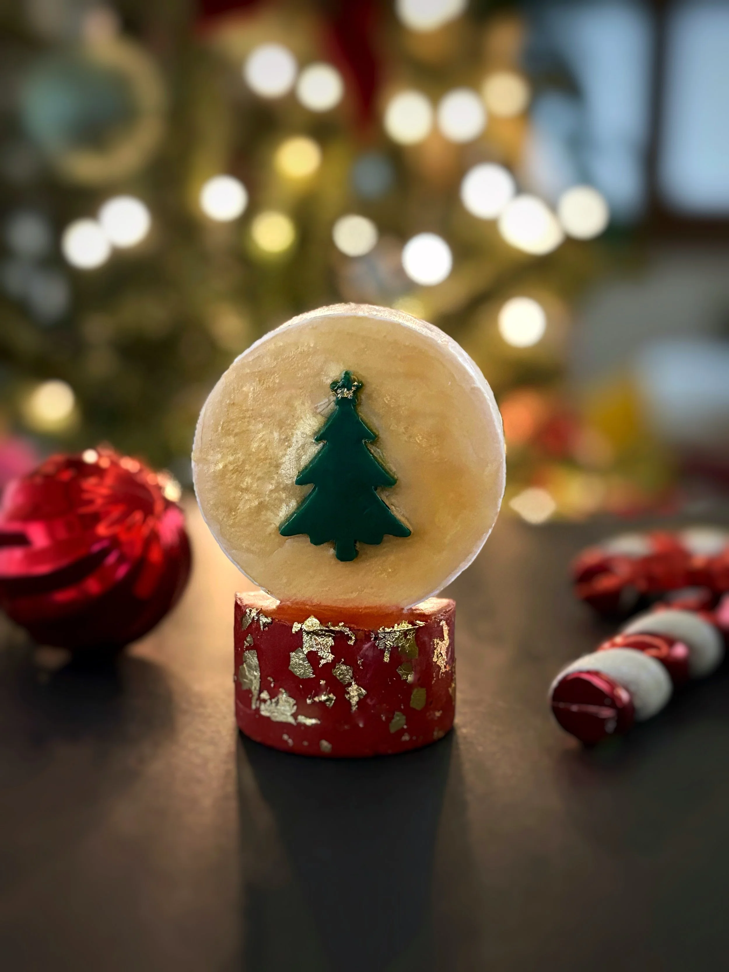 Decorative holiday Christmas scene with a candle featuring a green Christmas tree, Christmas ornaments, and a blurred Christmas tree with lights in the background.