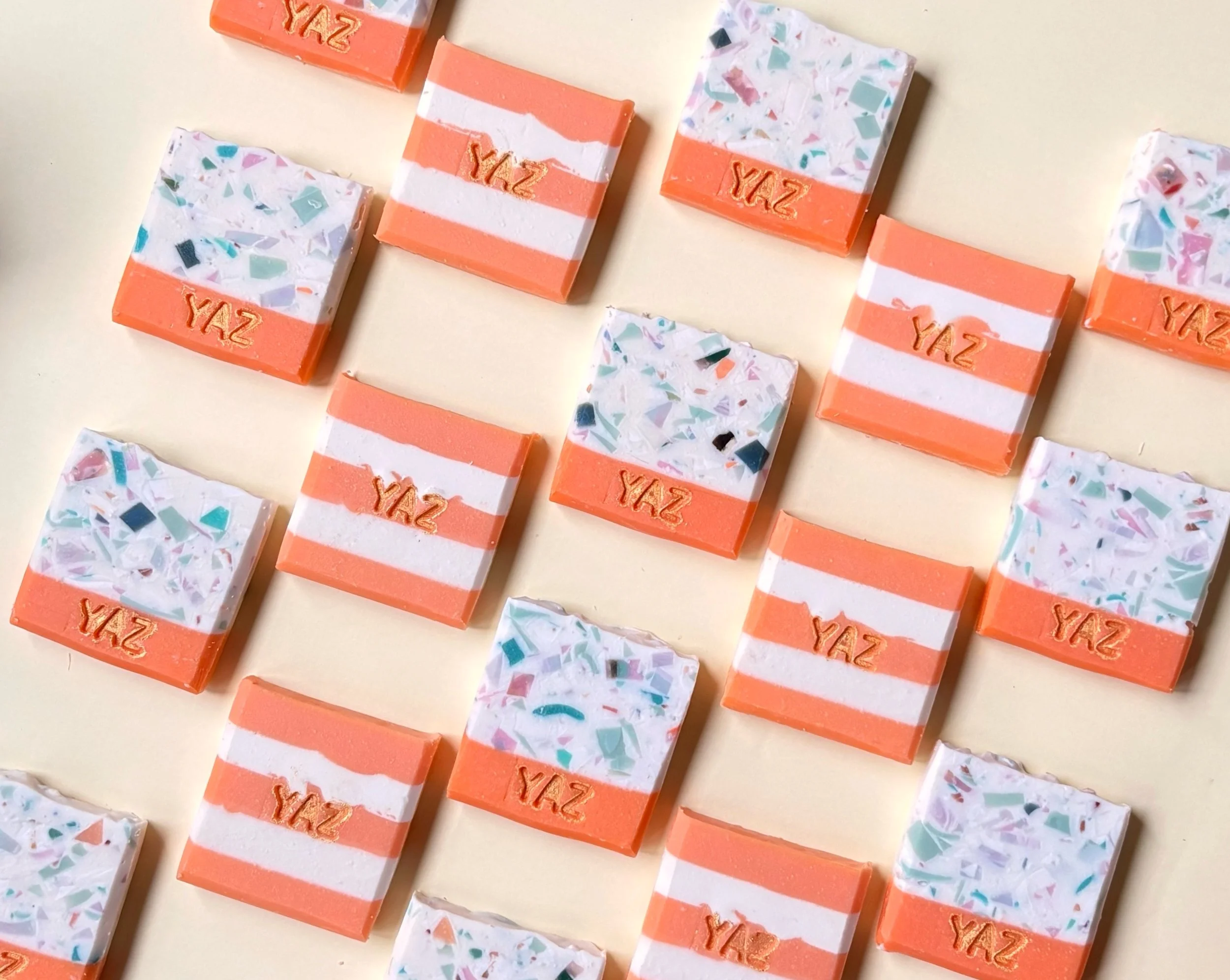 Flat lay of square patterned soaps with orange and white stripes and terrazzo designs, embossed with the word 'YAZ' on an off-white background.