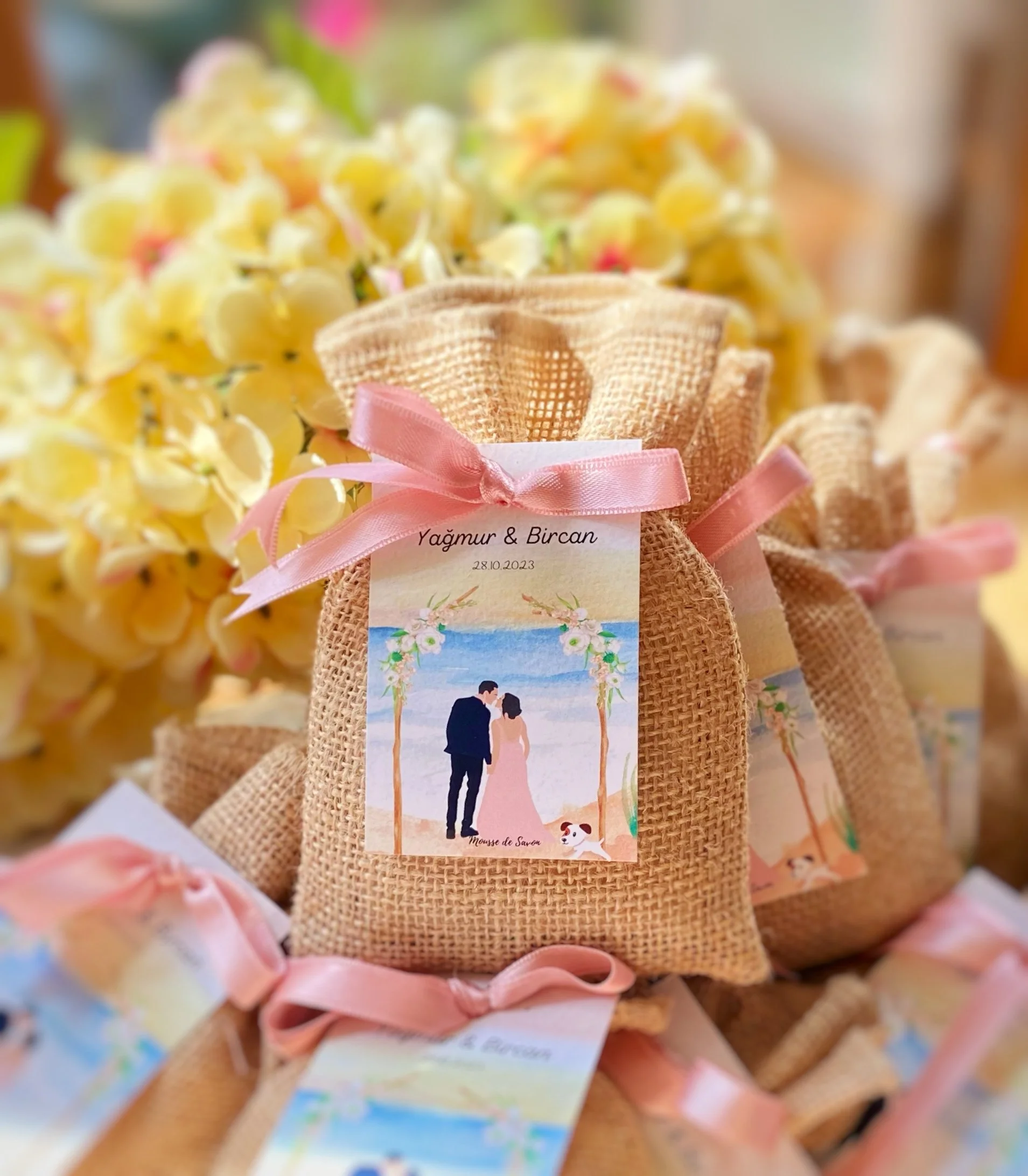 Small burlap gift bag decorated with pink ribbon, with a wedding-themed illustration and text, surrounded by yellow flowers.