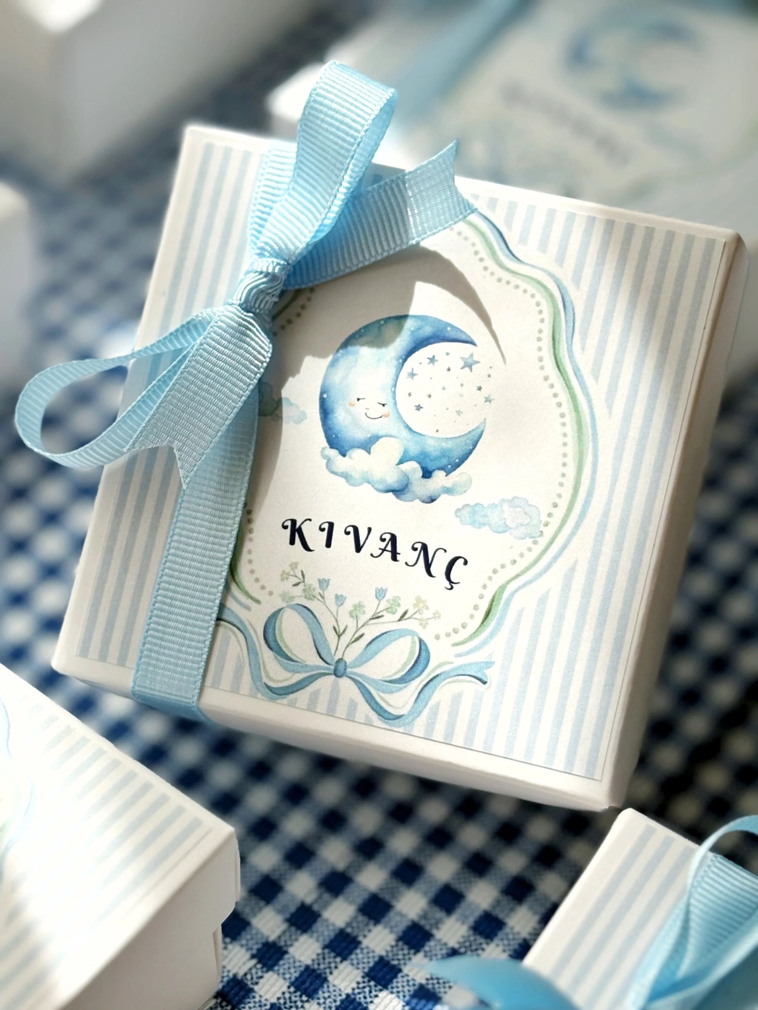 A baby shower favor box decorated with blue and white stripes, featuring a moon and star illustration, with a blue ribbon on top.