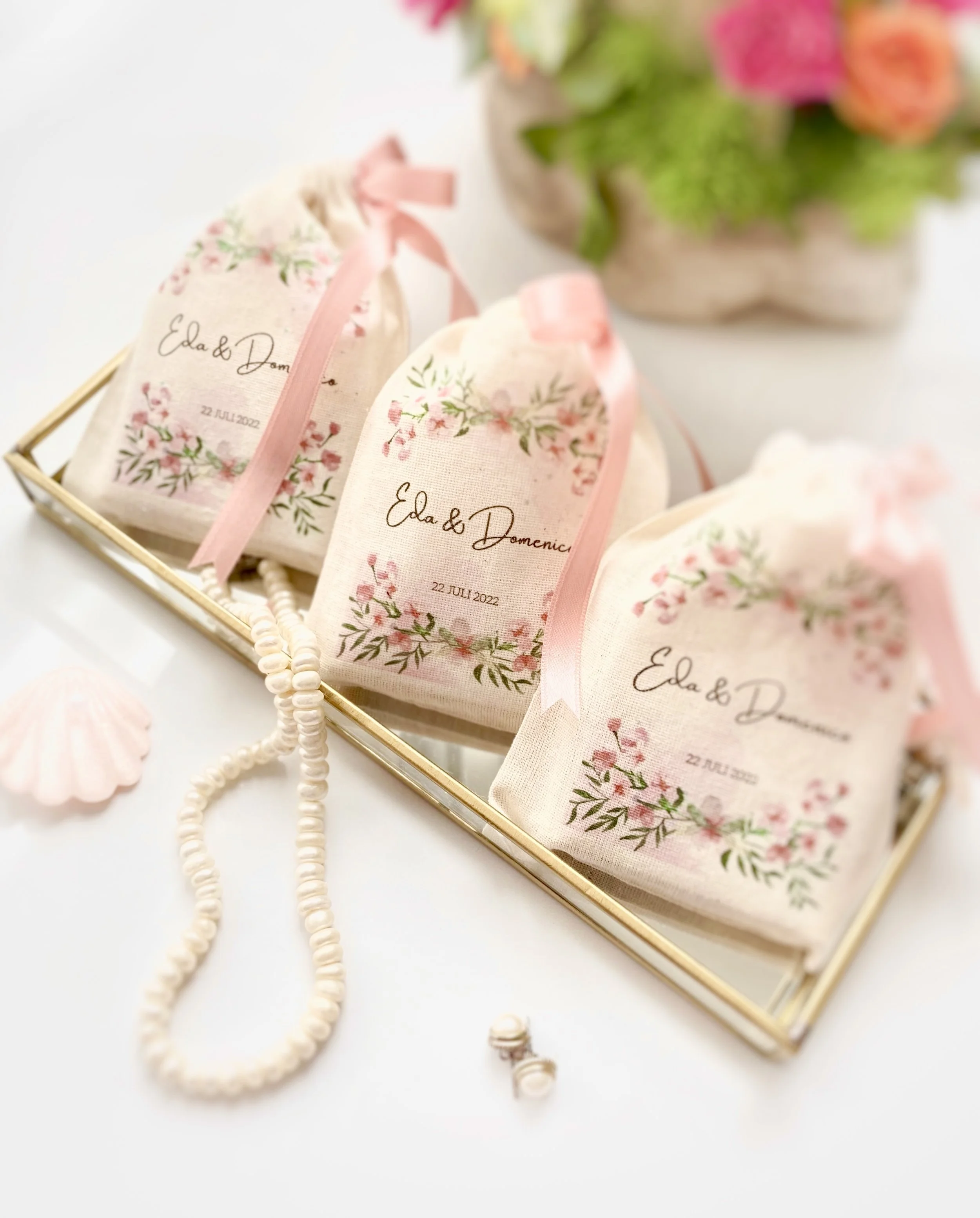 Three fabric pouches with floral designs and wedding date, placed in a golden tray, with pink ribbon ties. Also on the tray is a pearl necklace, a pair of pearl earrings, and a pink shell-shaped object, with a colorful flower arrangement in the backg