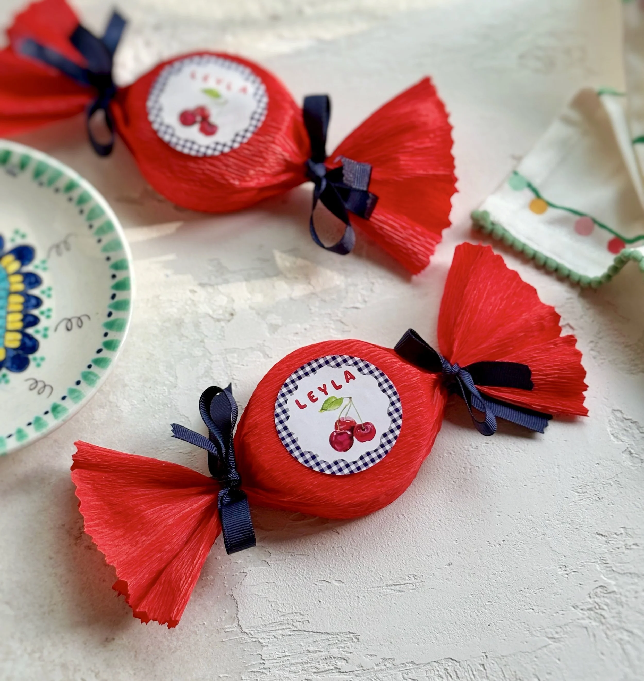 Two pieces of red wrapped candy with labels that say 'Leyla' and cherries, tied with black ribbons, surrounded by decorative ceramic dishware on a white textured surface.