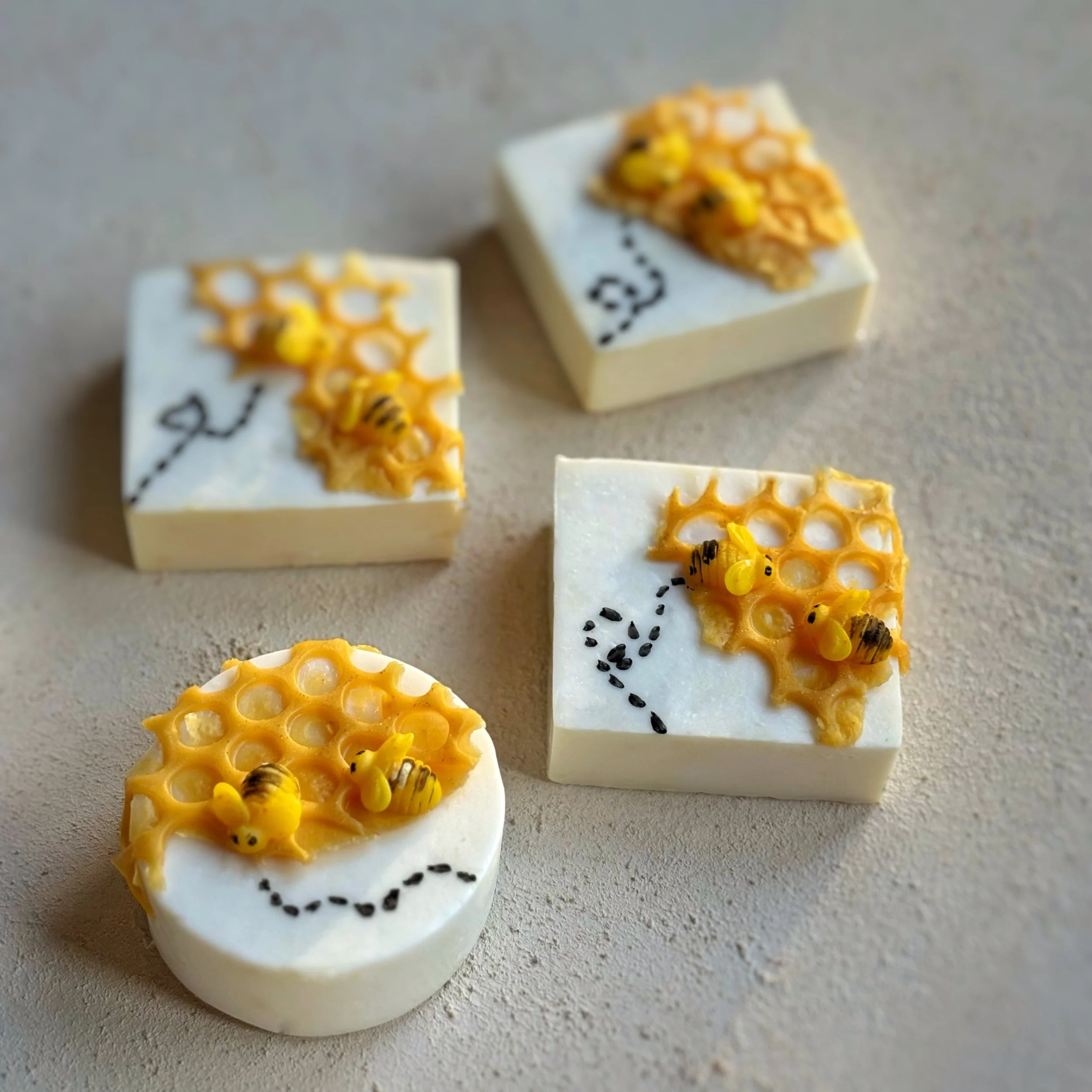Four small white soap confections decorated with honeycomb and tiny bee figures, arranged on a neutral surface.