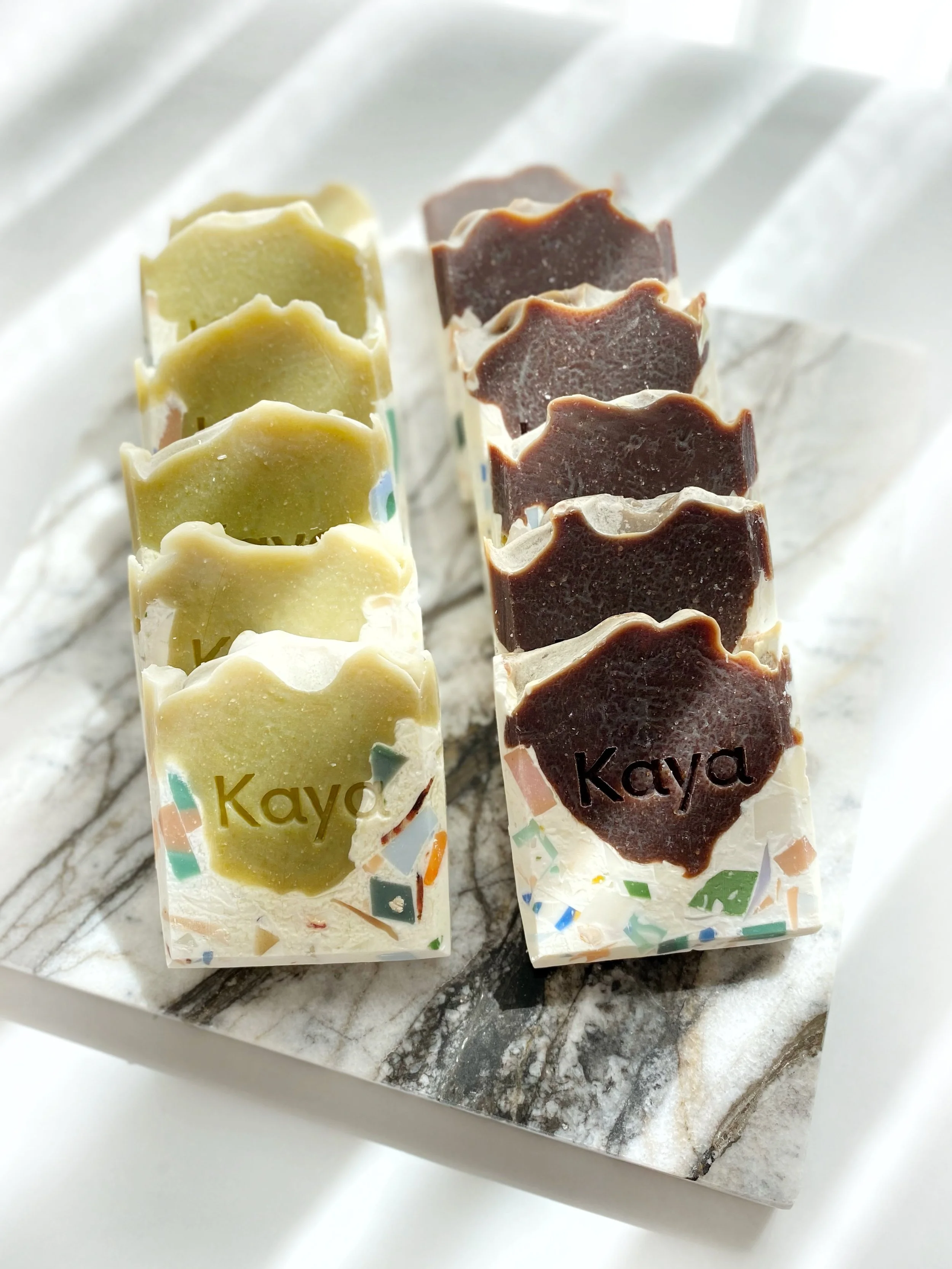 Two rows of soap bars, one with greenish soap and the other with dark brown soap, both with the word "Kaya" on them, arranged on a marble tray.