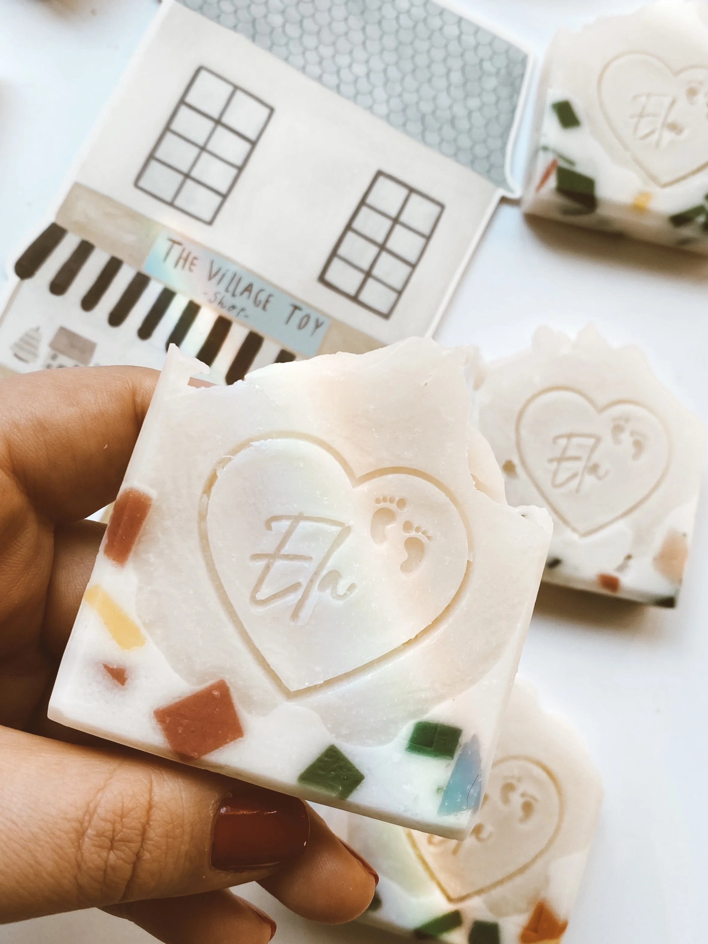 Hand holding a square bar of soap with a heart and footprint embossed on it, surrounded by other similar soaps, with a shop sign and packaging visible in the background.
