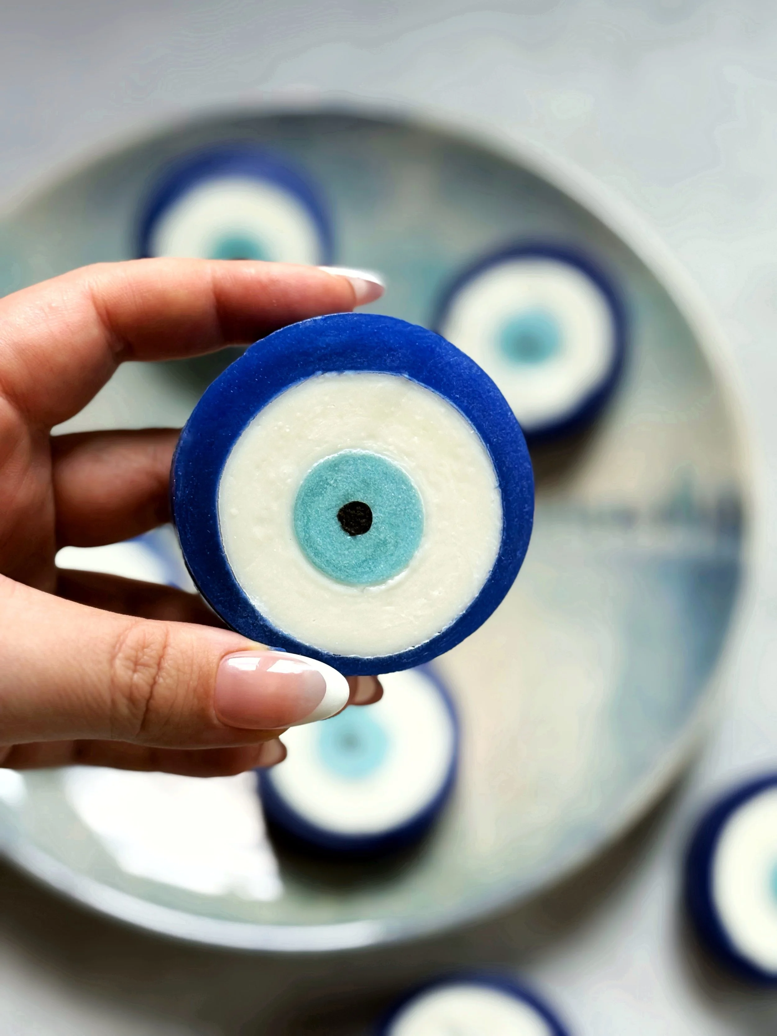 Hand holding a round cookie decorated like an evil eye with layers of blue, white, and light blue icing, on a white plate with more similar cookies in the background.