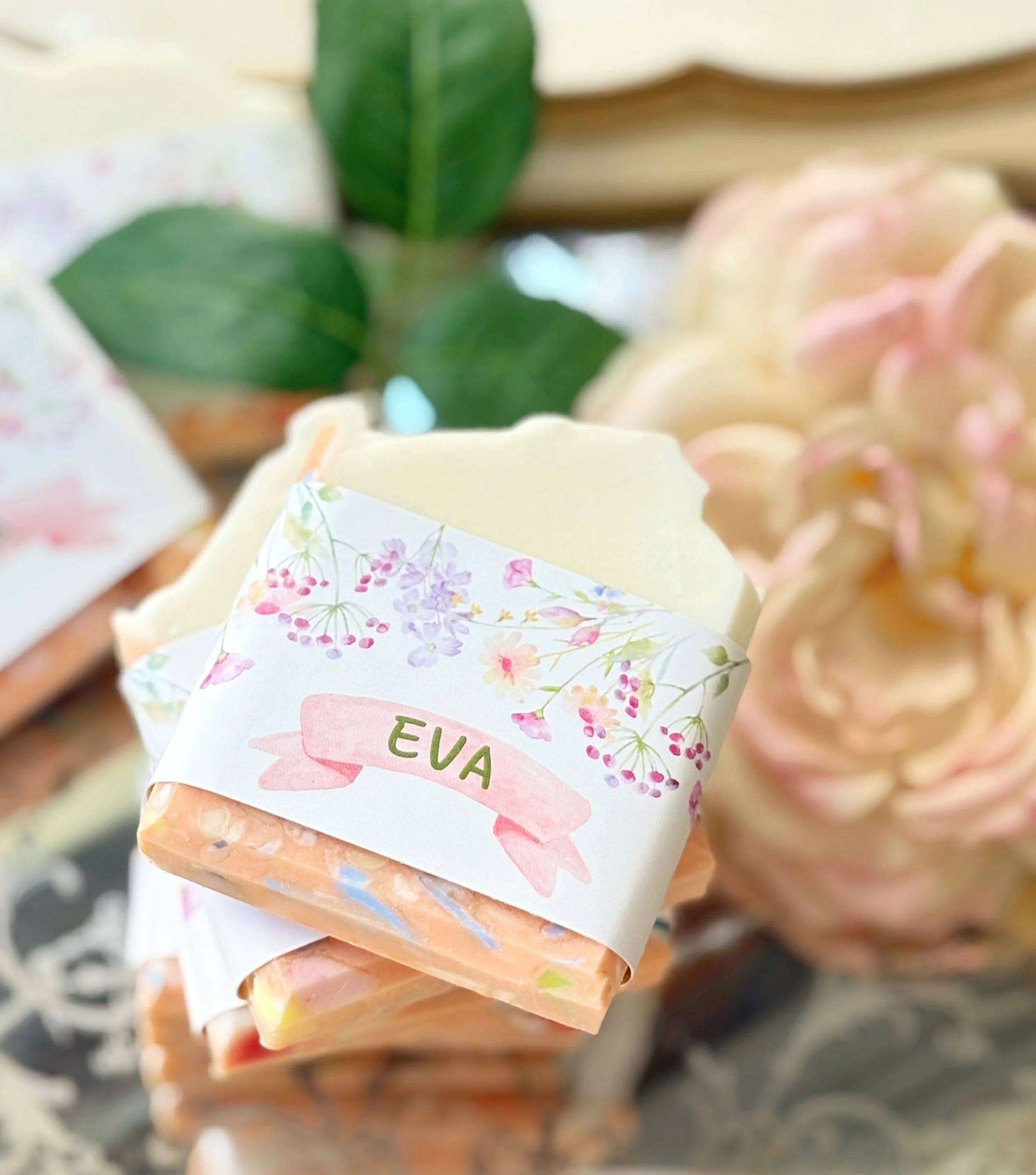 A soap bar wrapped with a floral label reading "EVA," placed among decorative flowers and green leaves.