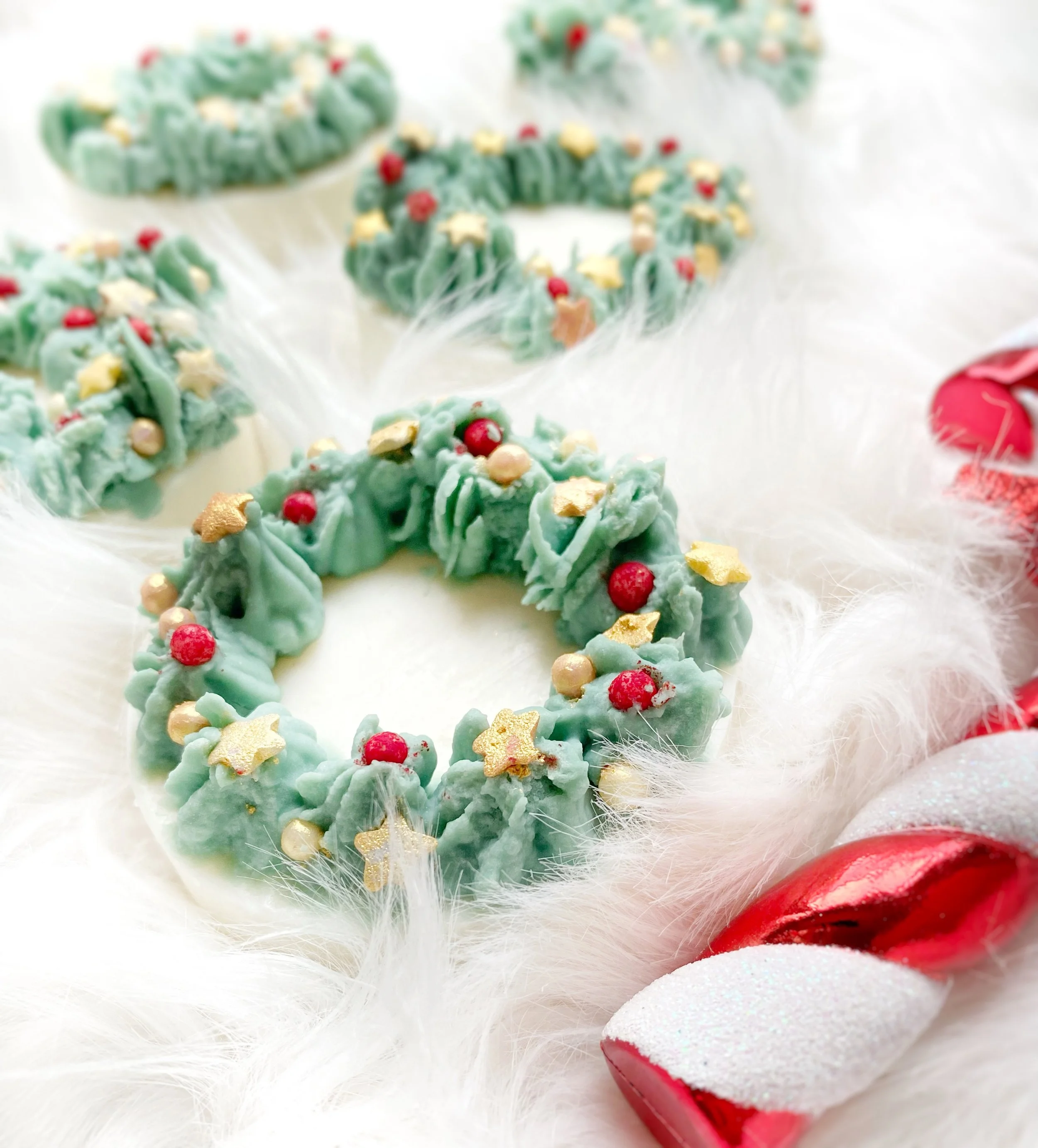 A close-up of decorative Christmas wreath soaps with green icing, red and gold edible beads, on a white furry surface, with a red and white peppermint candy near.