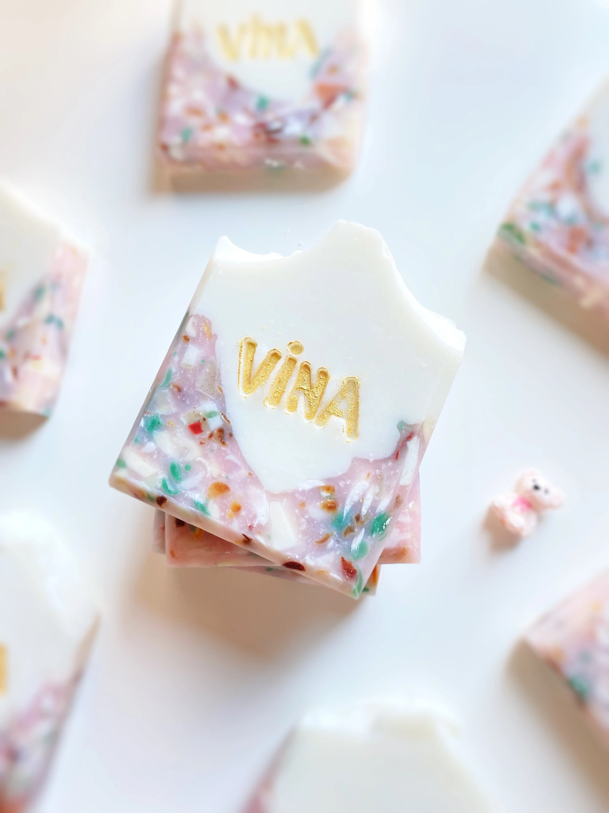 Bar of soap with the name 'VINA' embossed in gold, decorated with colorful sprinkles, on a white surface with additional soap bars around it.