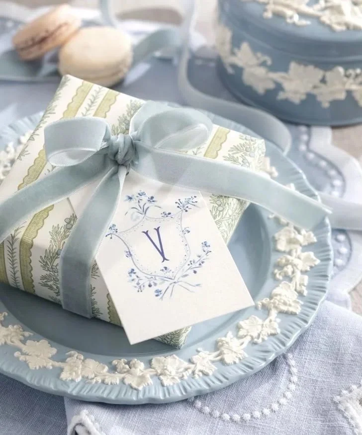 A gift box wrapped in patterned paper with a white and blue design, tied with a pale blue ribbon, placed on a light blue plate with lace detailing, on a table with decorative items.
