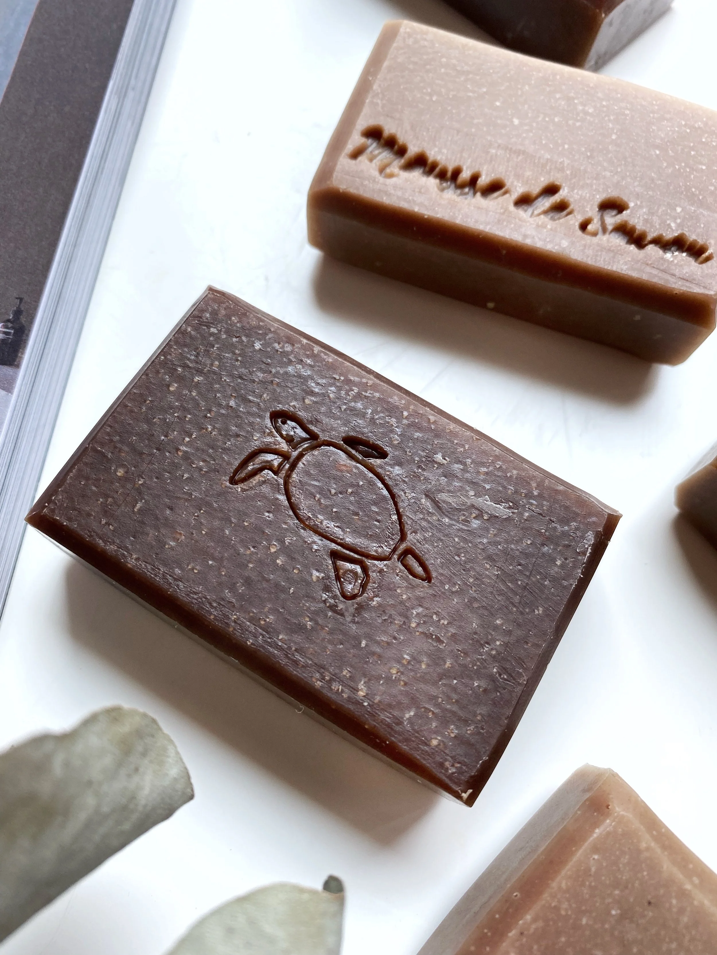 Bars of handmade soap with carved and embossed designs, including a turtle and a phrase, on a white surface.