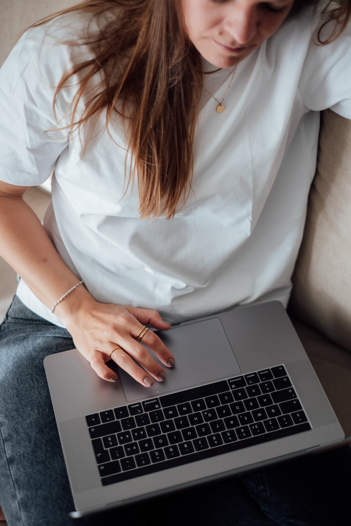 Frau mit braunen Haaren sitzt auf einem Sofa, trägt ein weißes T-Shirt und benutzt einen Laptop.