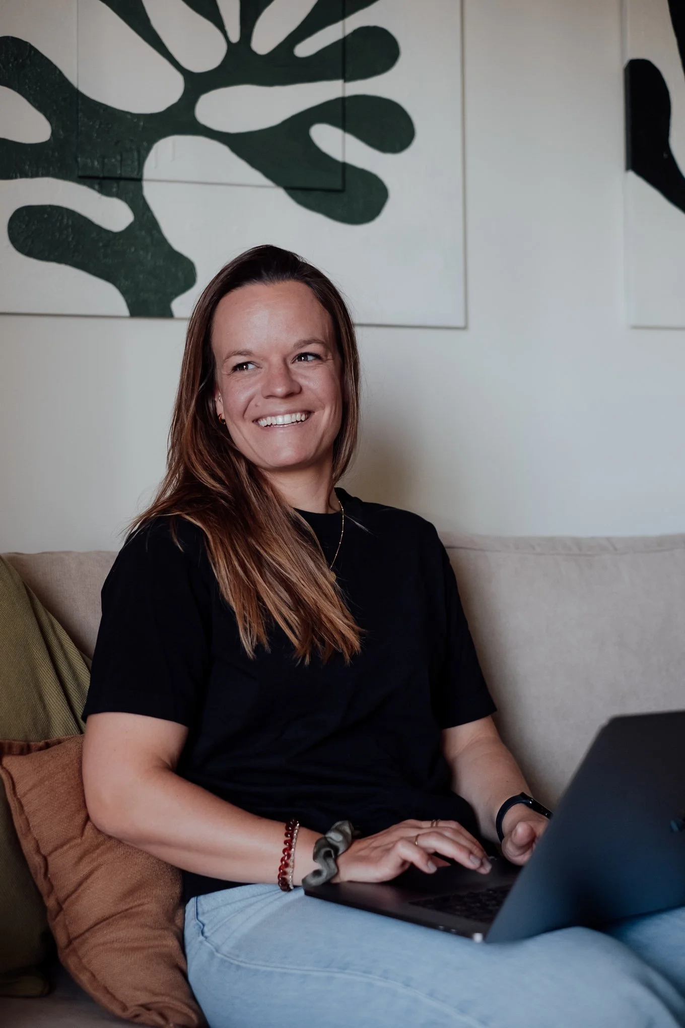 Lächelnde Frau sitzt auf einem Sofa mit Laptop auf ihrem Schoß, hinter ihr grüne und weiße Wandkunst