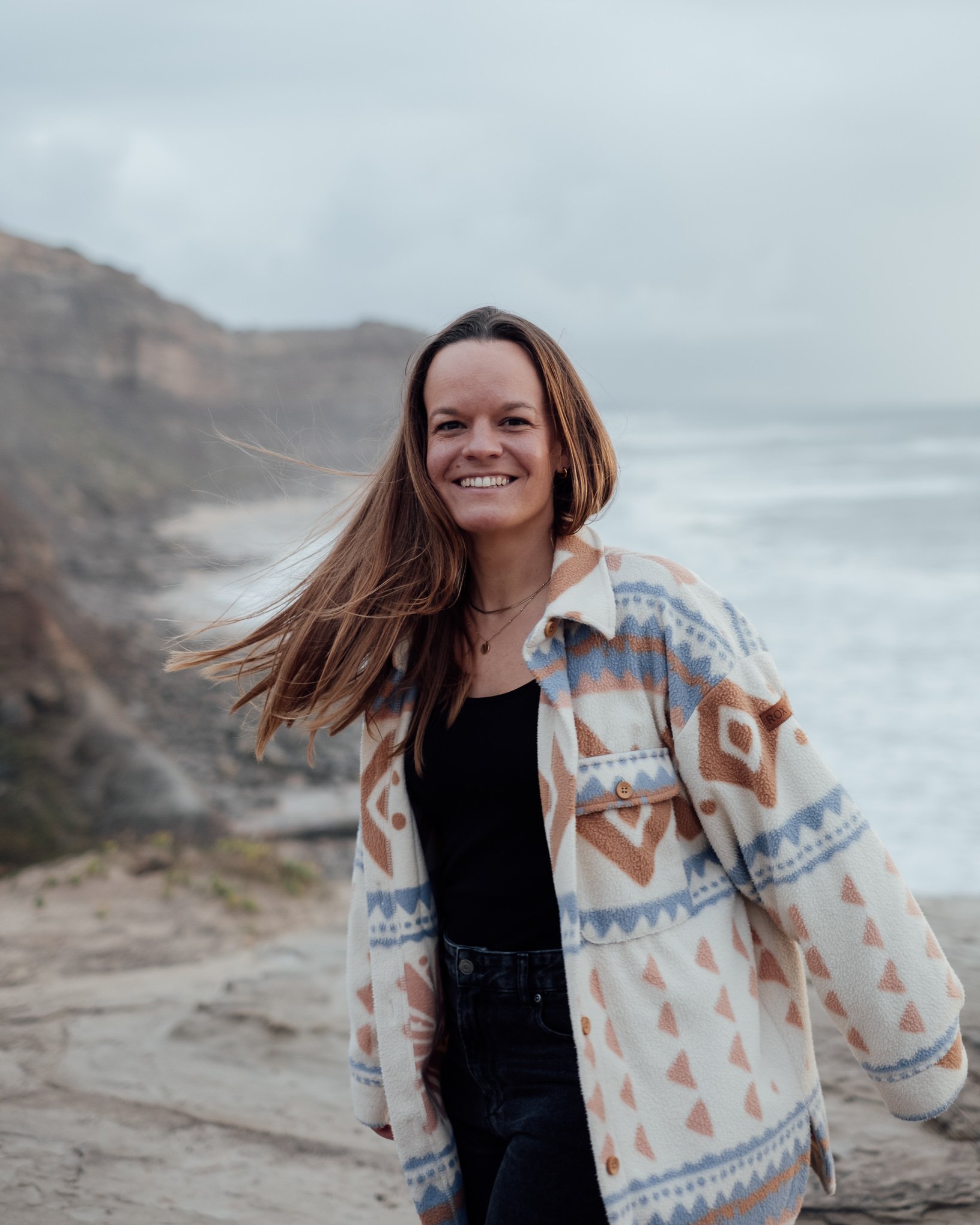 Eine lachende junge Frau steht an einem Strand mit Felsen und Meer im Hintergrund, trägt eine bunte, gemusterte Jacke und ein schwarzes Oberteil.