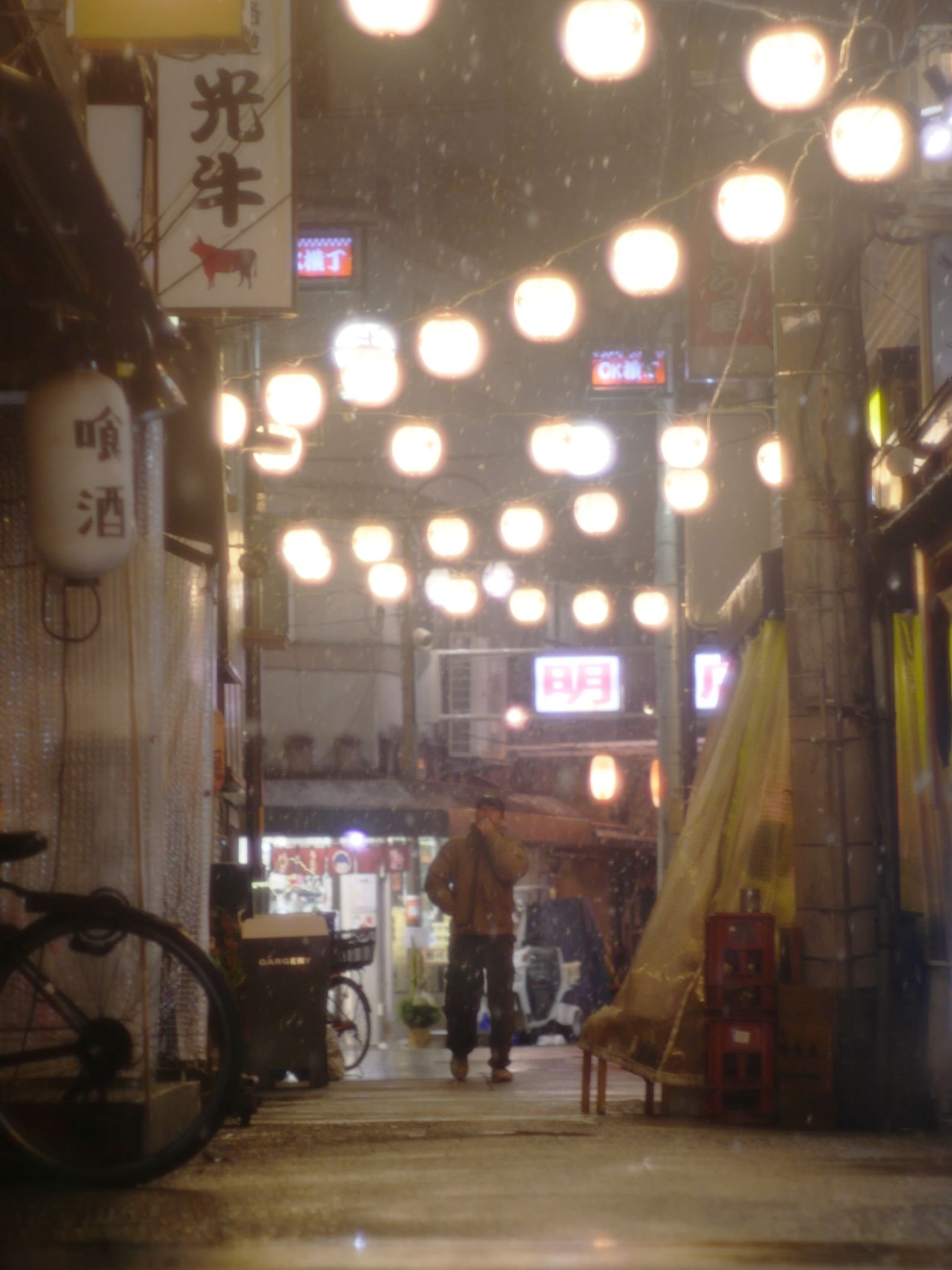 雨の夜の小さな飲み屋街、暖かい光の提灯や電飾、傘をさす男性と自転車が見える。