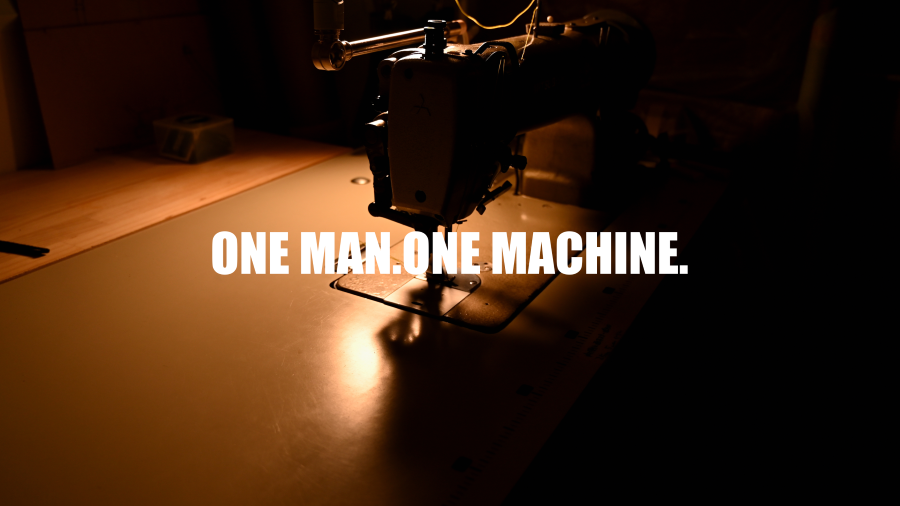 縫製機械と木製の作業台、暗い工房の中にある光の当たる部分に文字『ONE MAN. ONE MACHINE.』