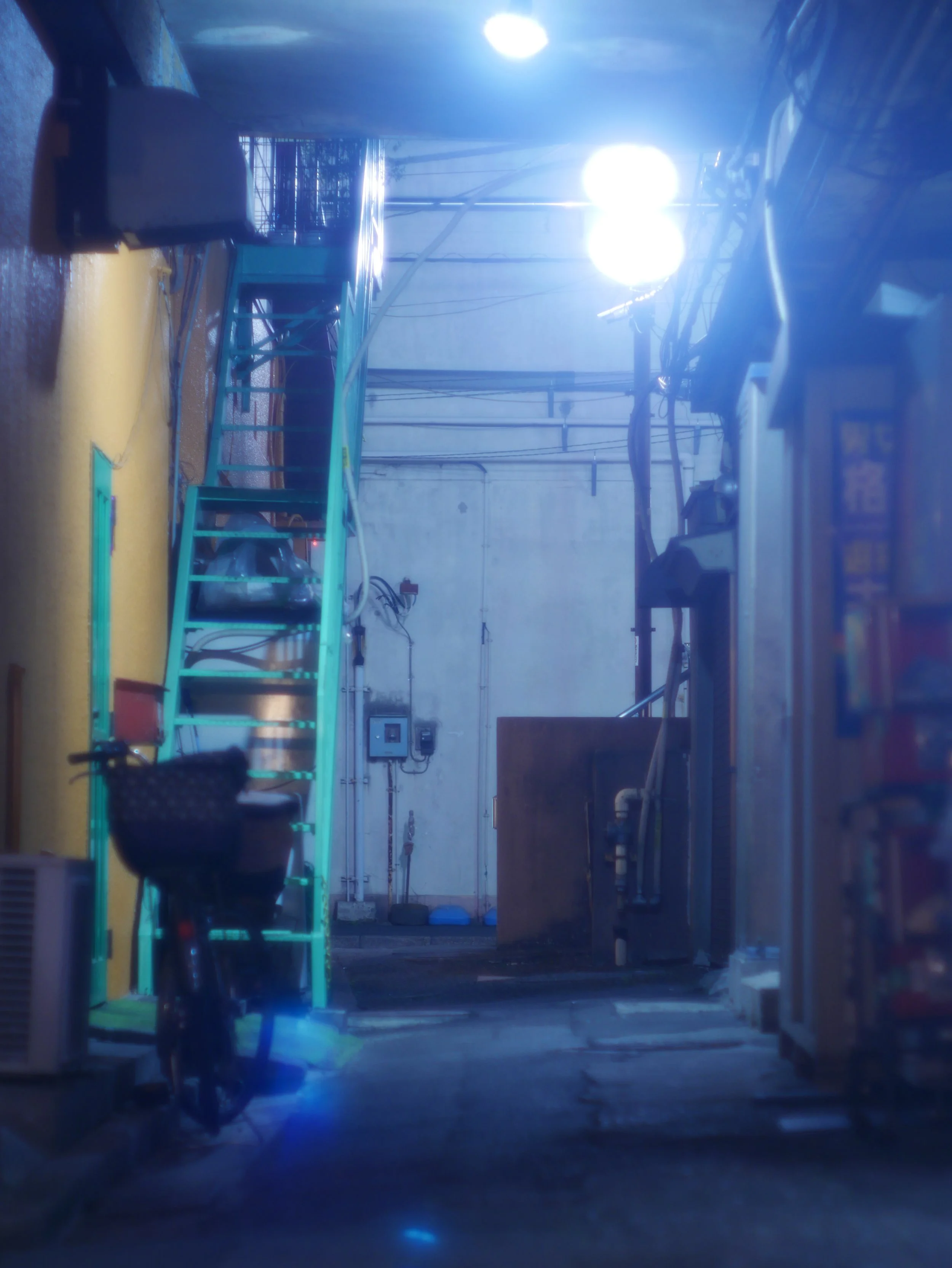 狭い路地、左側に自転車、青い階段、電線と電柱、背景に白い壁と明かり。