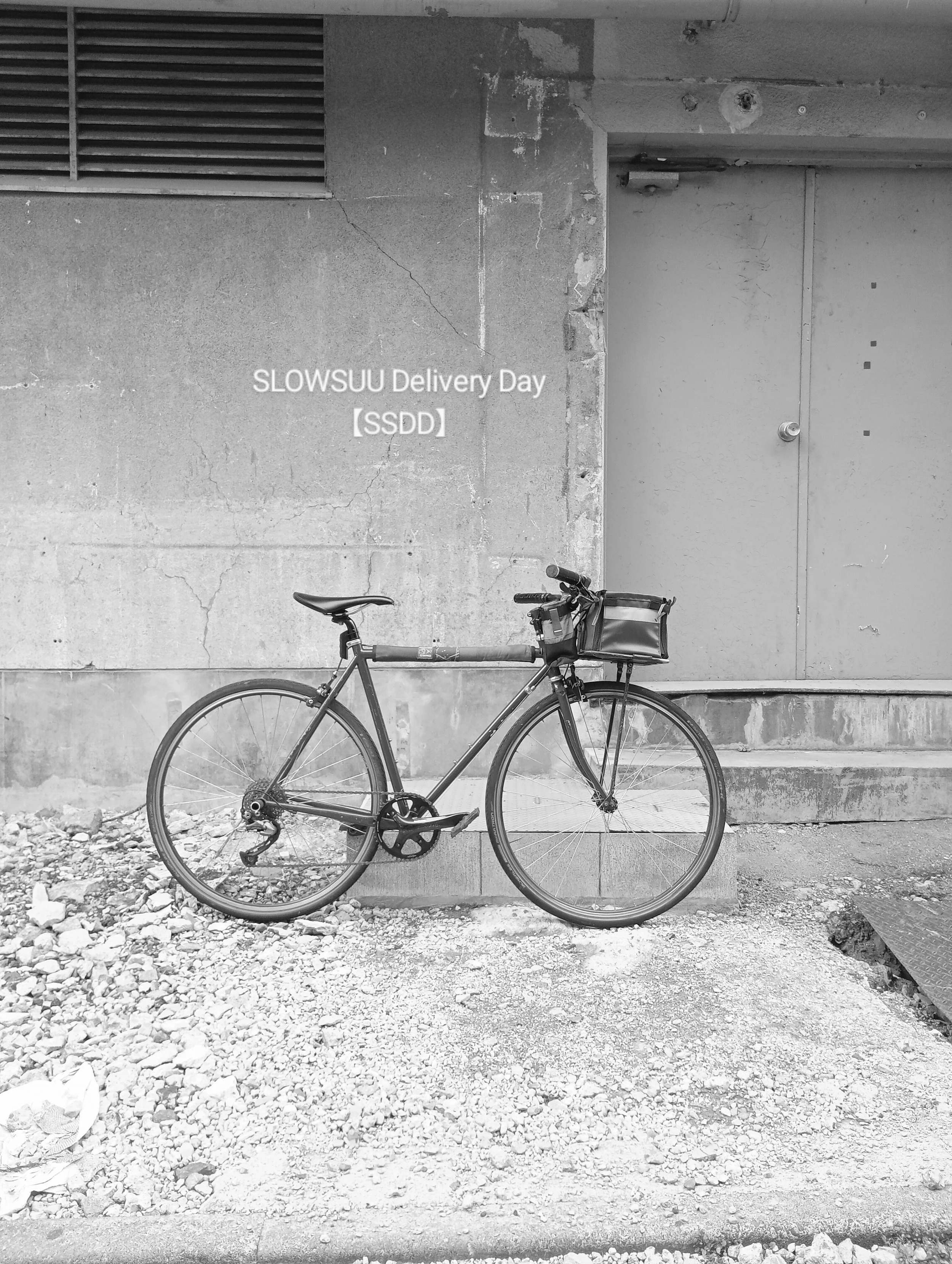 白黒の自転車がコンクリートの道に止まっている。背景に壁とドアがある。壁に「SLOWSUU Delivery Day 【SSDD】」と書かれている。