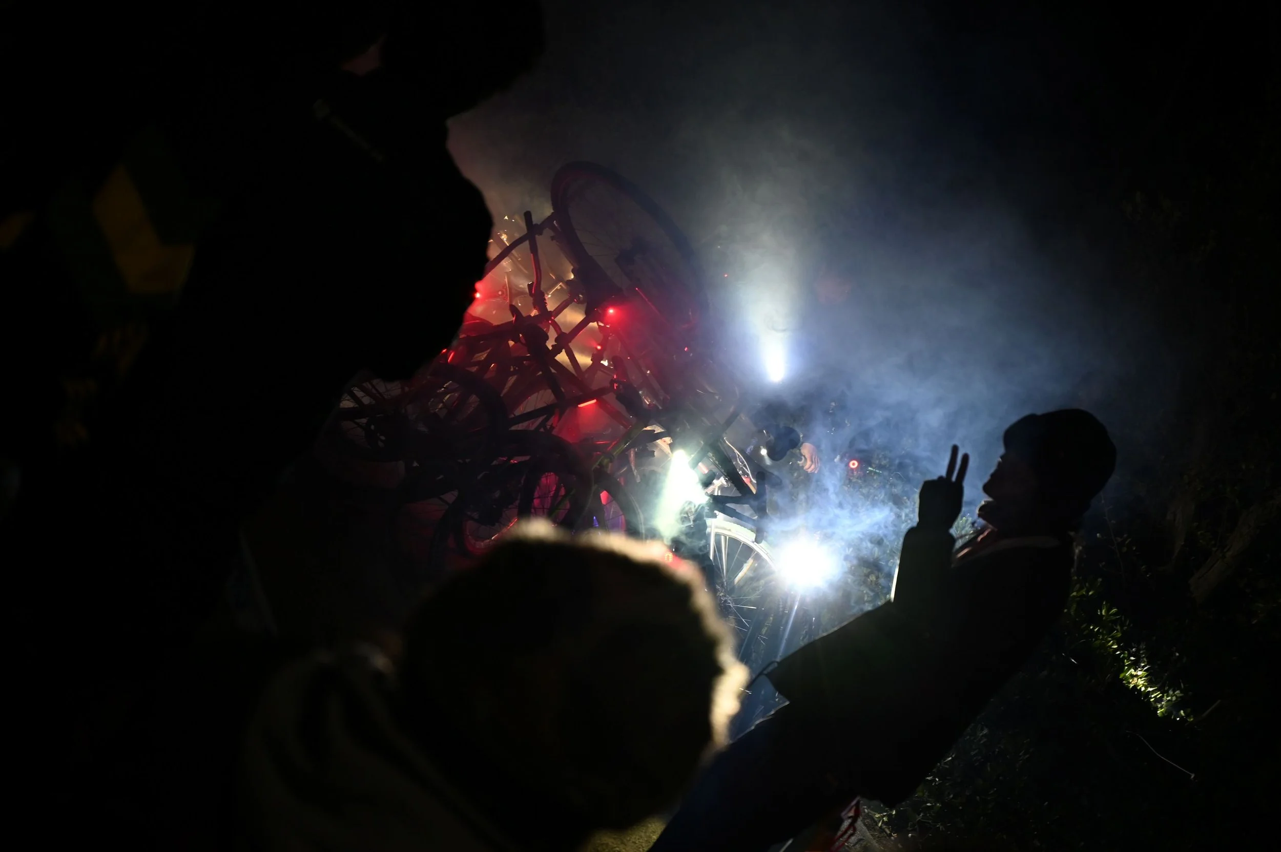 夜間に自転車が倒れ、数人の人が集まっている。背景に煙と光があり、事故や修理の瞬間を捉えているようだ。