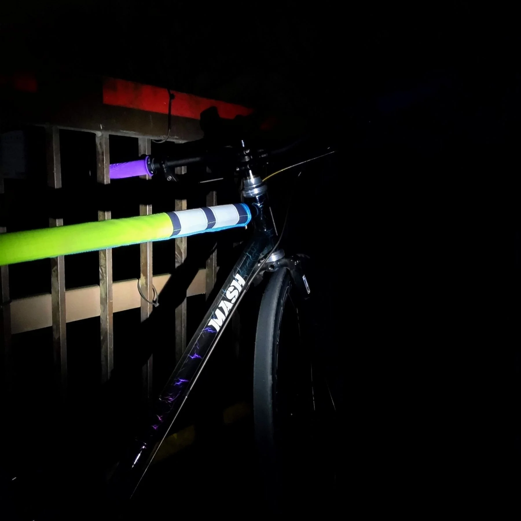 暗闇の中にある自転車の部分的なアップ。青と紫のライトが当たっており、背後には木製のフェンスが見える。