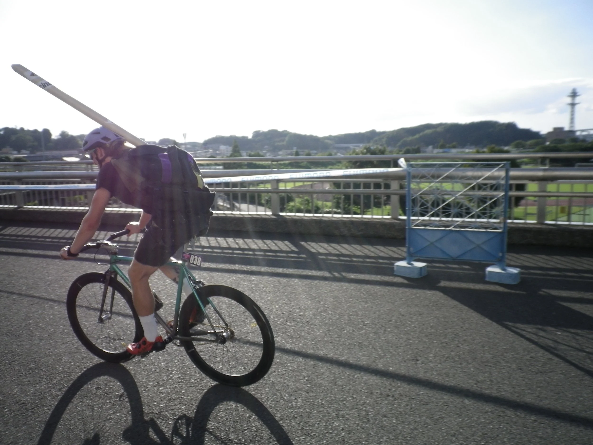 ヘルメットをかぶった男性がロードバイクで走っている。背中にリュックを背負っている。道路の横にフェンスがあり、その向こうに緑の風景と遠くに展望台が見える。