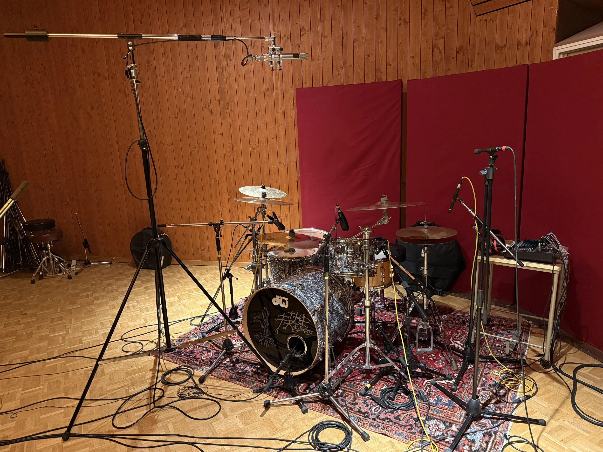 Ein professionelles Aufnahme- oder Tonstudio mit einem Schlagzeug in der Mitte, umgeben von Mikrofonen und Kabeln, in einem Raum mit Holzverkleidung an der Wand und roten Schallabsorbern.