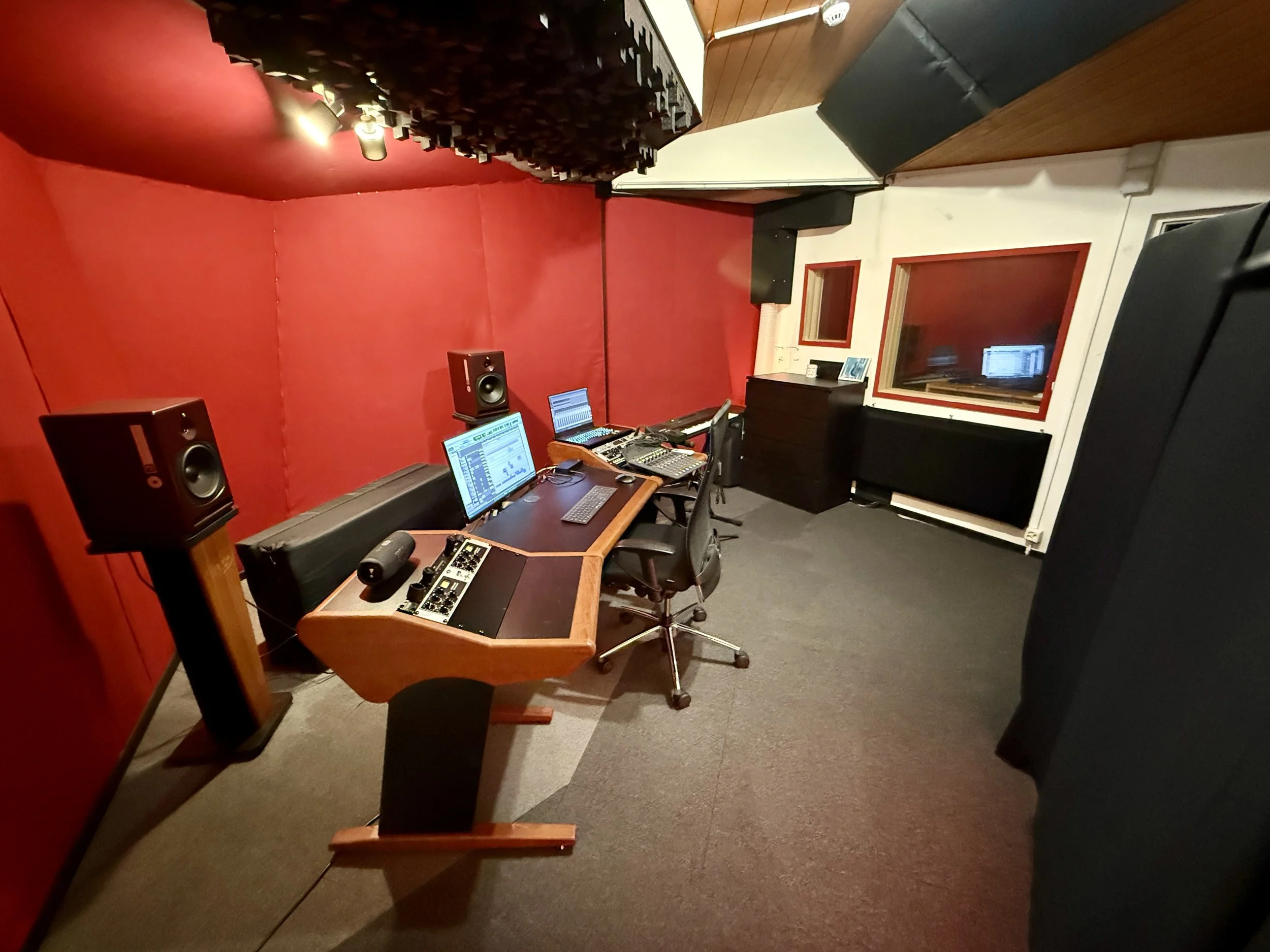 Aufnahme- oder Sprechraum mit Musik- und Aufnahmeausrüstung, roten Wandpaneelen, Fenster, Schreibtisch, Stuhl und Lautsprechern.