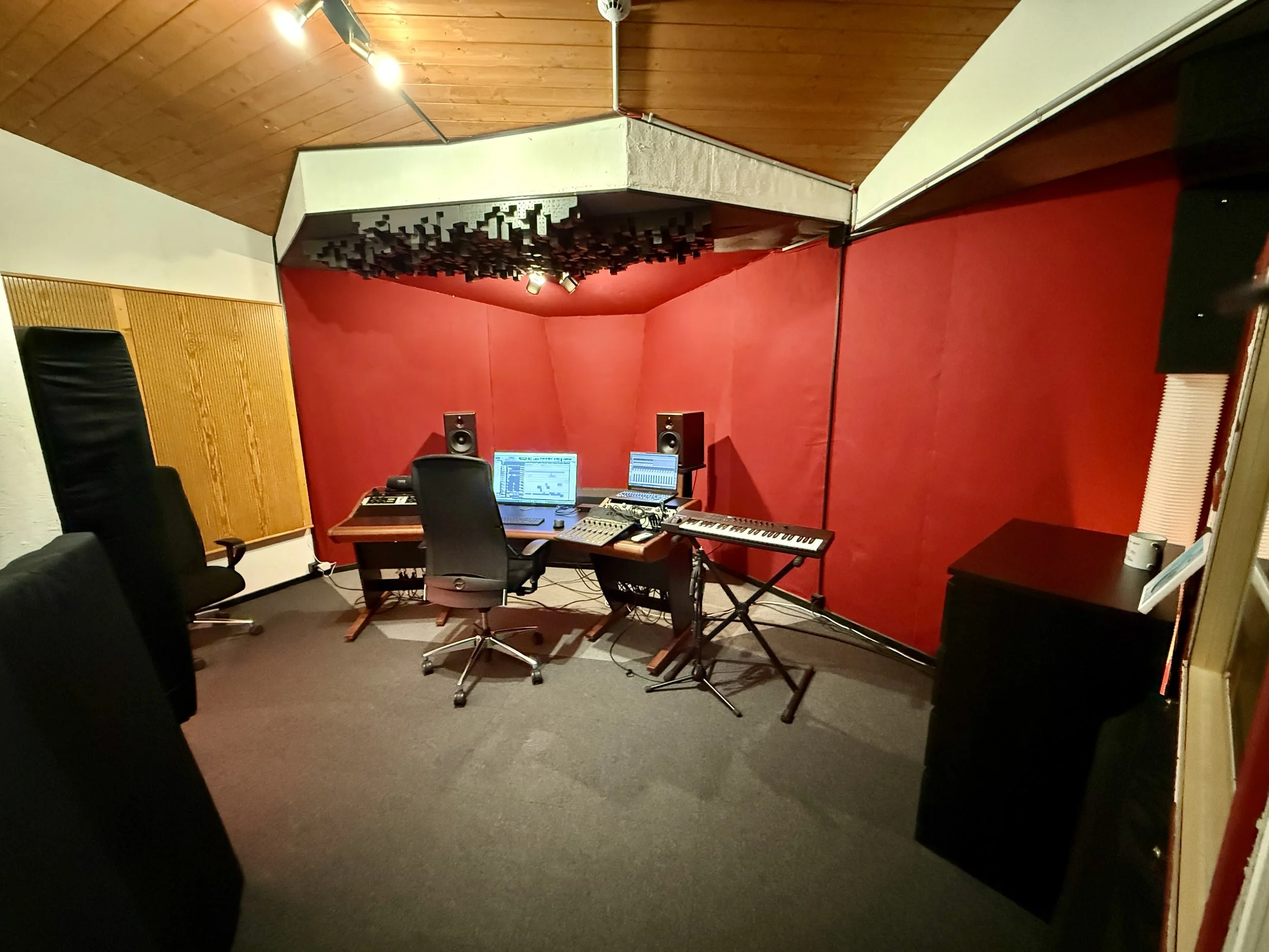 Aufnahmen eines Musikproduktionsraums mit Schreibtisch, Computer, Studiomonitore, Keyboard und Aufnahmeequipment, rote Wandverkleidung, akustische Deckenabsorber.