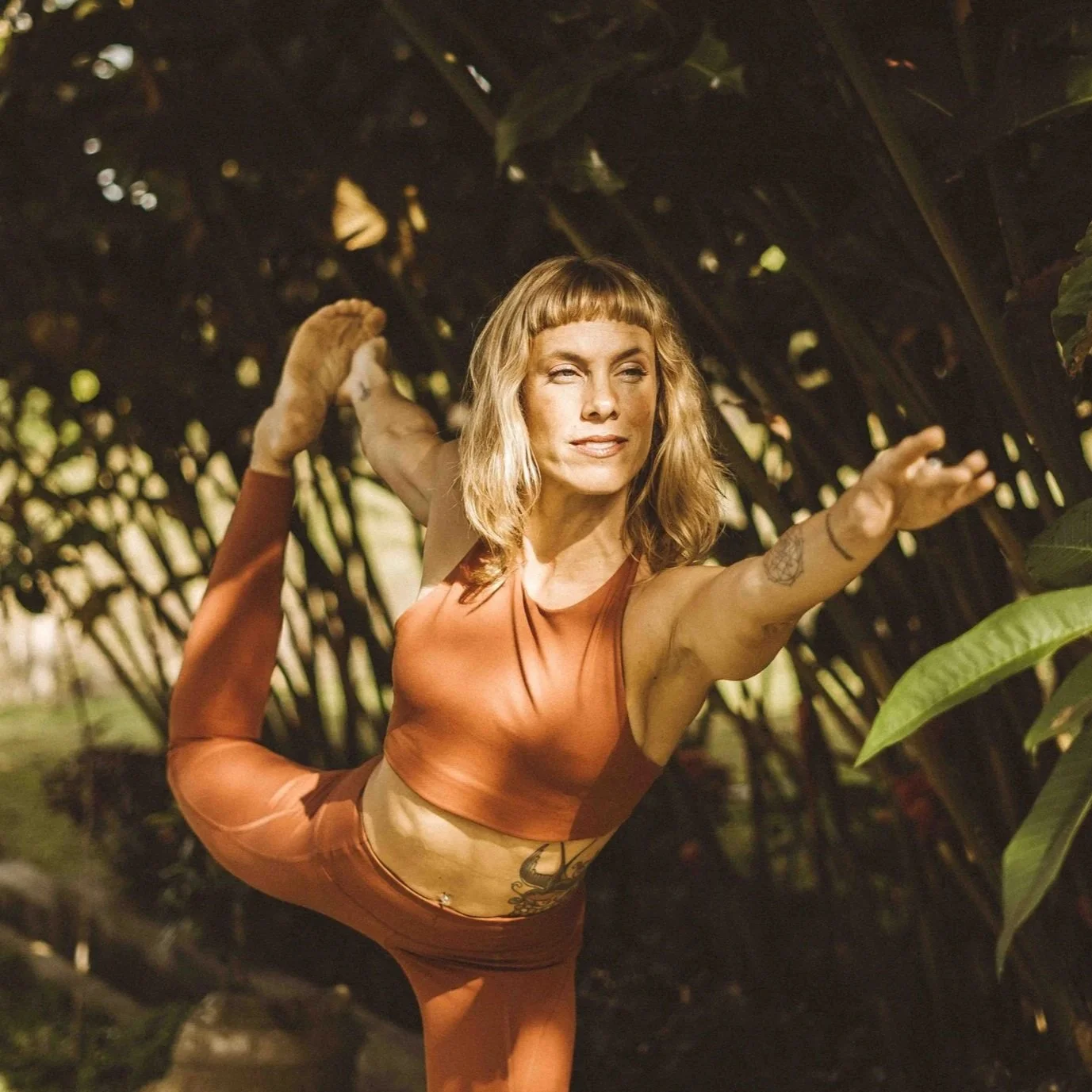 A woman practicing yoga outdoors, performing a standing backbend pose amidst lush greenery and large leaves.