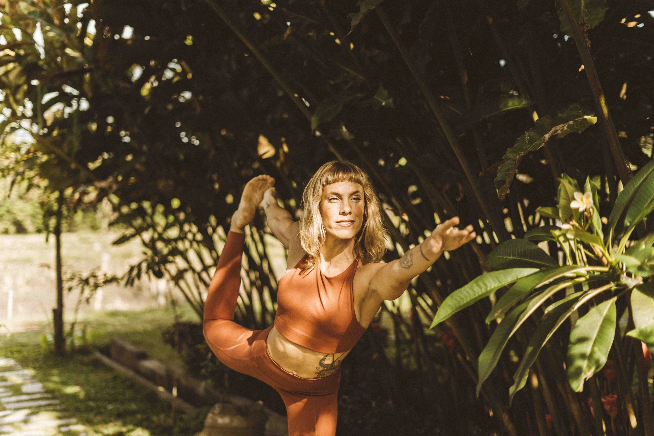 A woman practicing yoga outdoors under a large leafy tree, posed in a balancing pose with one leg bent behind her and one arm extended forward.