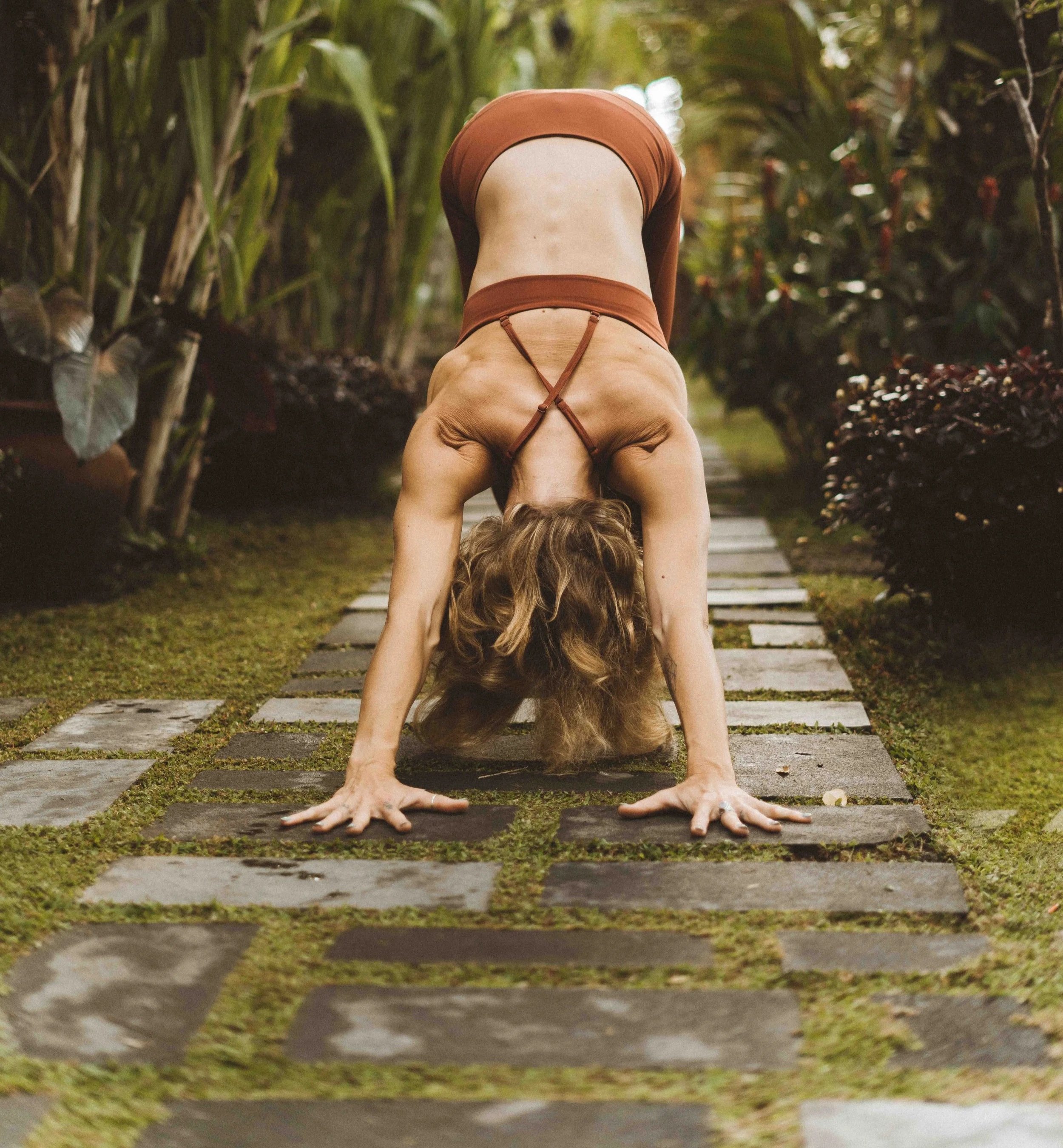 A woman practicing yoga outdoors on a stone path, performing a forward bend with her hands on the ground and her head hanging down, surrounded by lush greenery.
