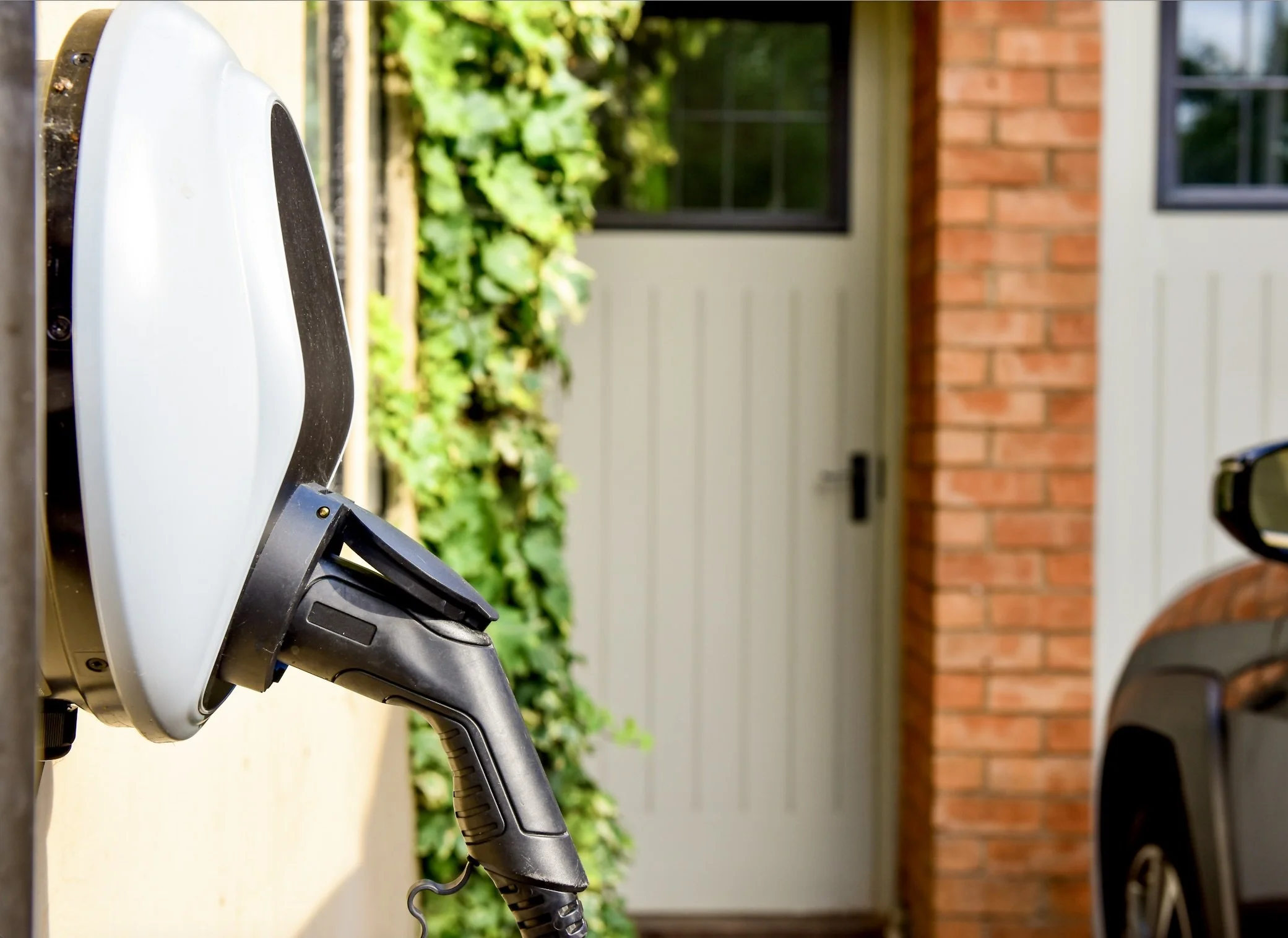 Electric vehicle charging station with a cable plugged in, situated outdoors next to a brick house wall and a garage door, with greenery visible in the background.