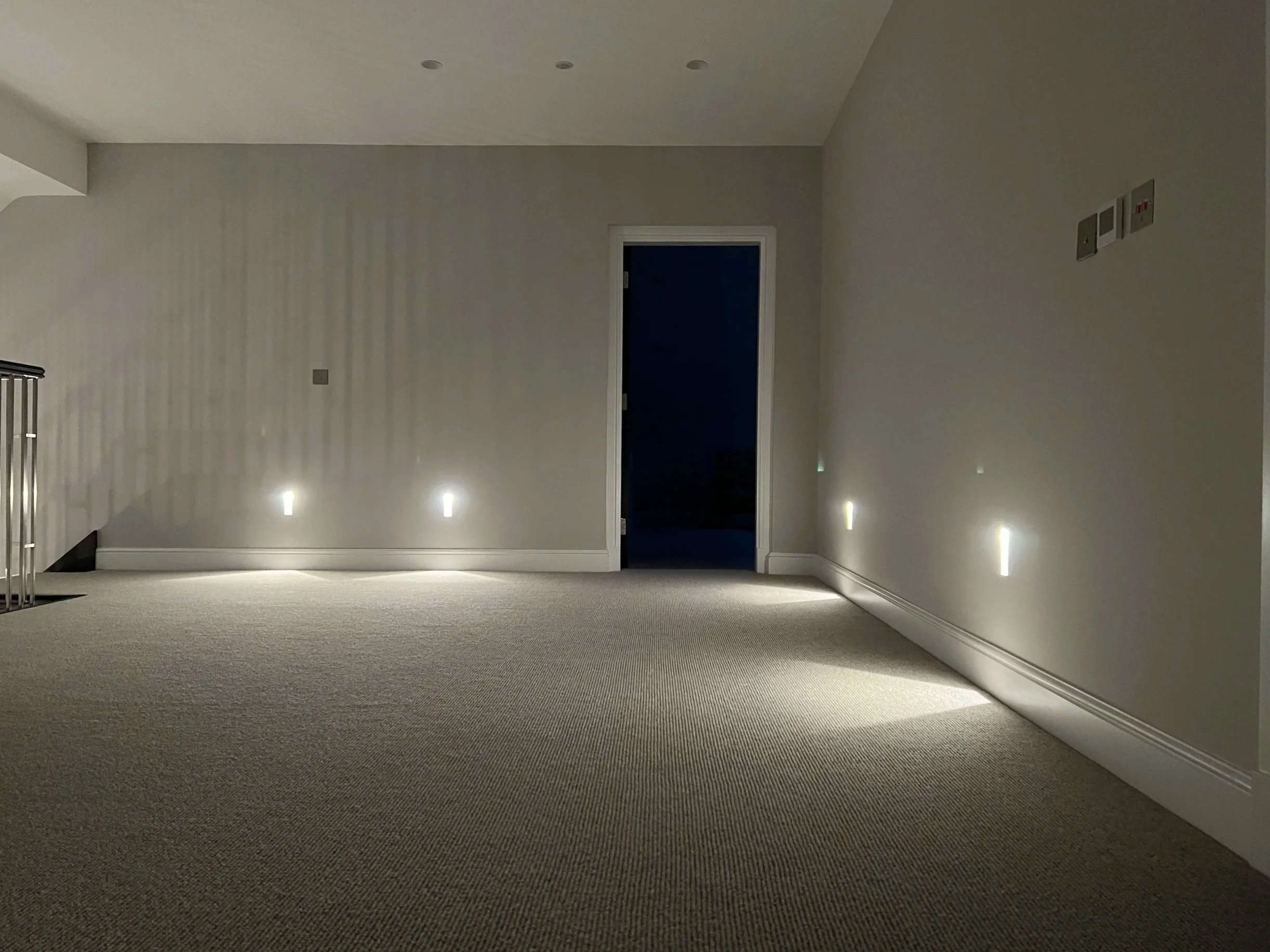 Empty room with wall-mounted lights casting shadows, carpeted floor, and a door at the end of the room.