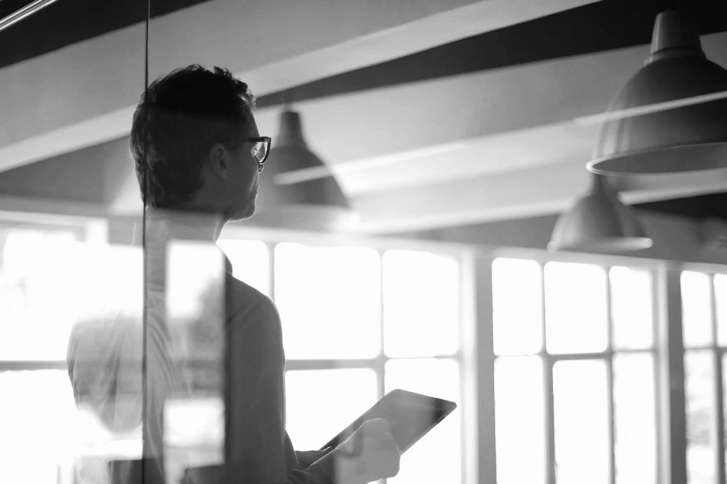 A person with glasses and short hair, seen in silhouette, standing indoors and looking at a tablet device, with large windows and hanging lights in the background.