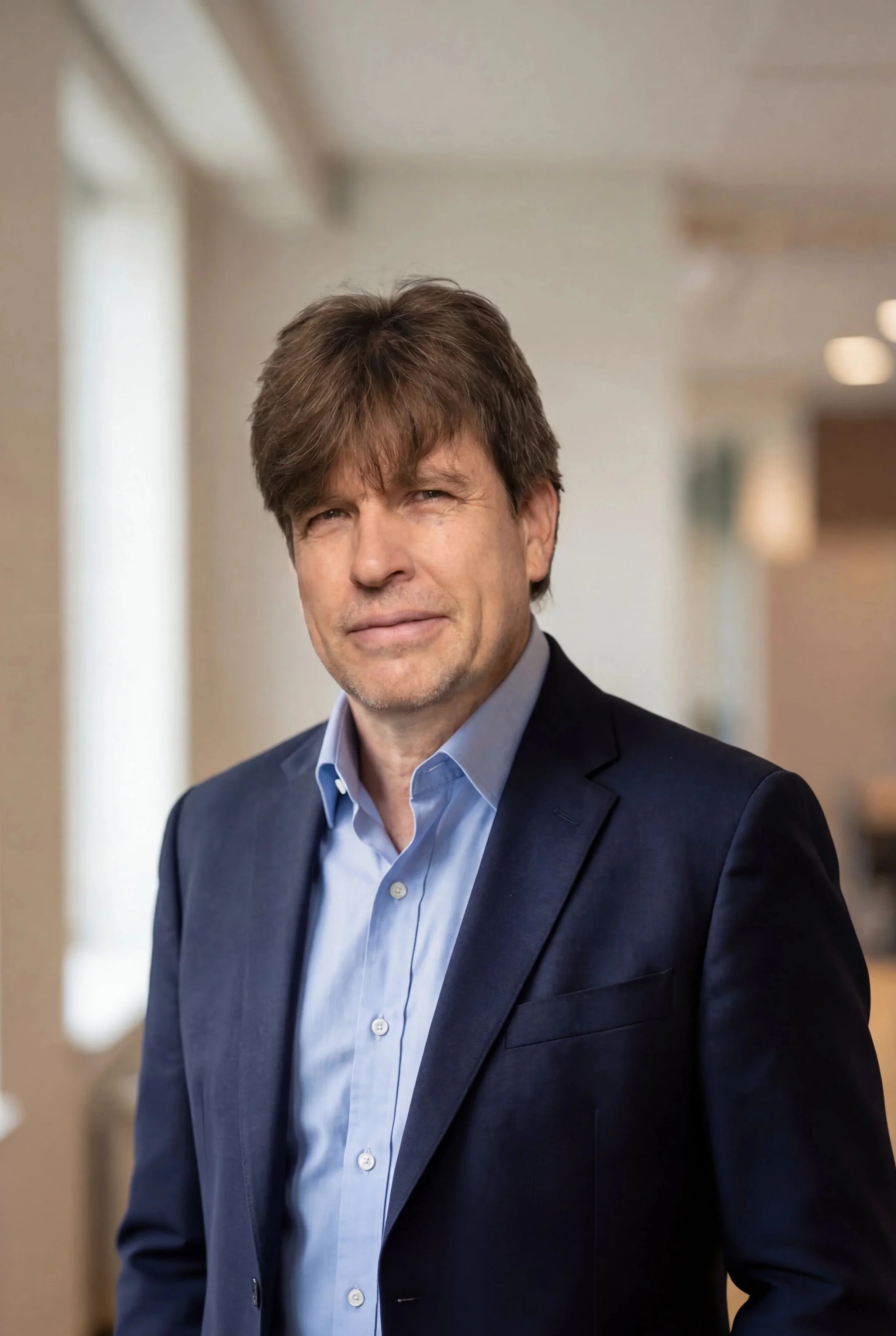 A middle-aged man with brown hair wearing a dark blue suit and light blue dress shirt, standing indoors with blurred background.