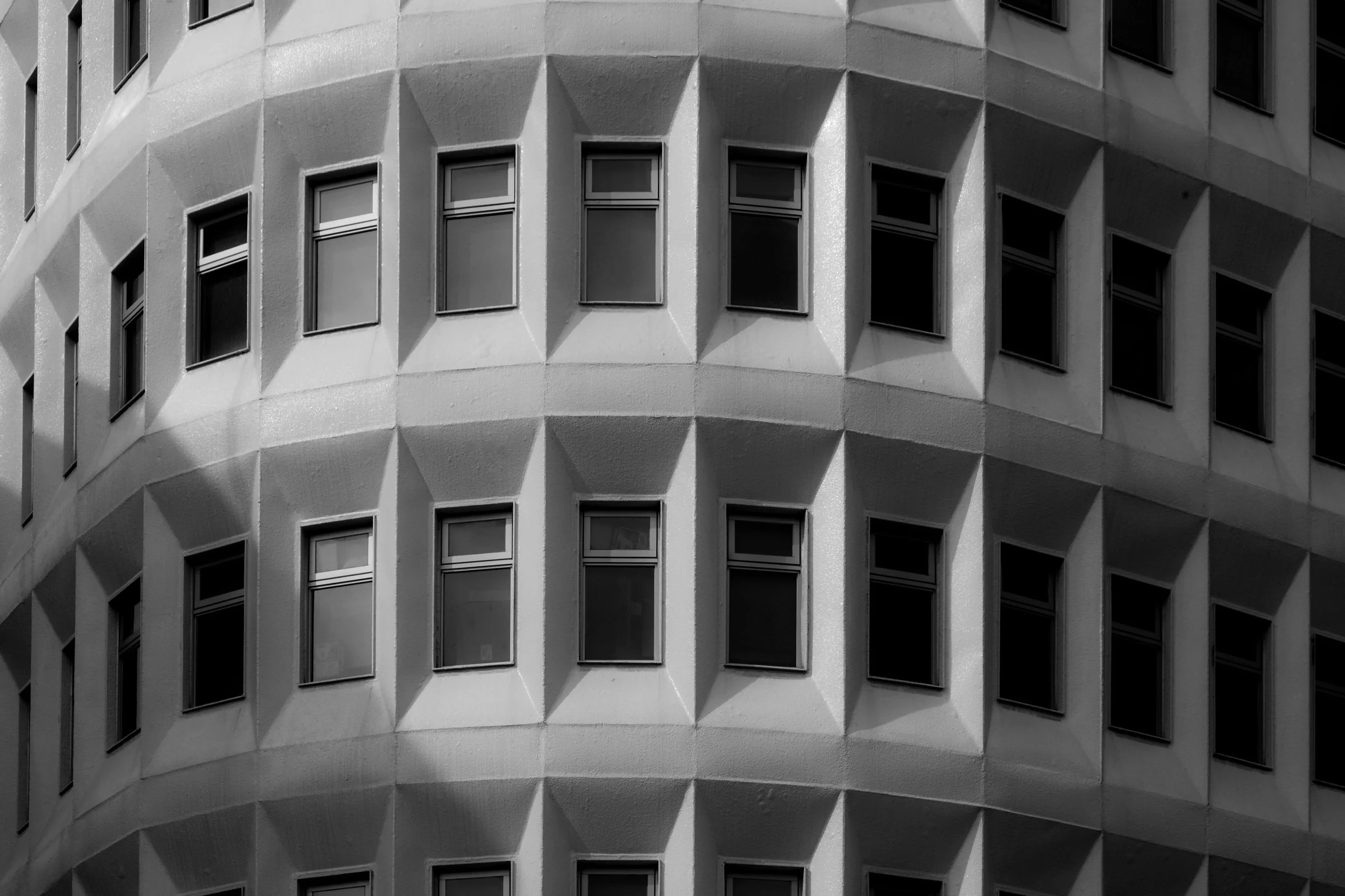 Close-up of a modern building facade with multiple rectangular windows, featuring geometric patterns created by protruding sections of the wall, in black and white.