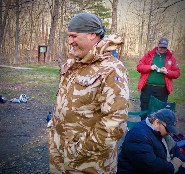 A man smiling outdoors in a wooded area, wearing a tan camouflage jacket with a UK flag patch, a gray headscarf, and hands in his pockets, with two other people in the background, one with a red jacket and cap and another in a blue jacket and glasses.