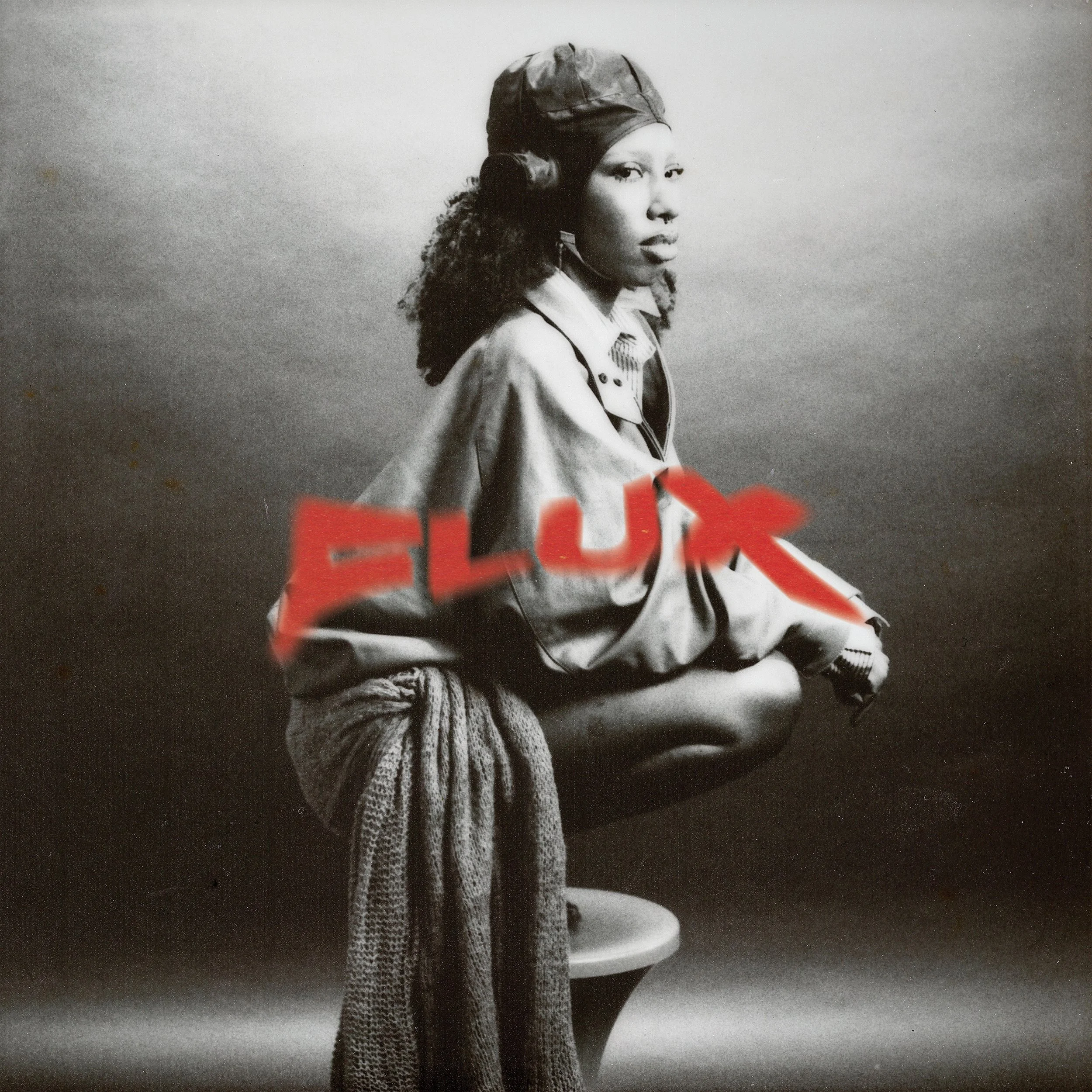Black and white photo of a woman squatting on a stool, wearing a jacket, a hat, and a knitted scarf. The word 'FLUX' is written in red across the image.