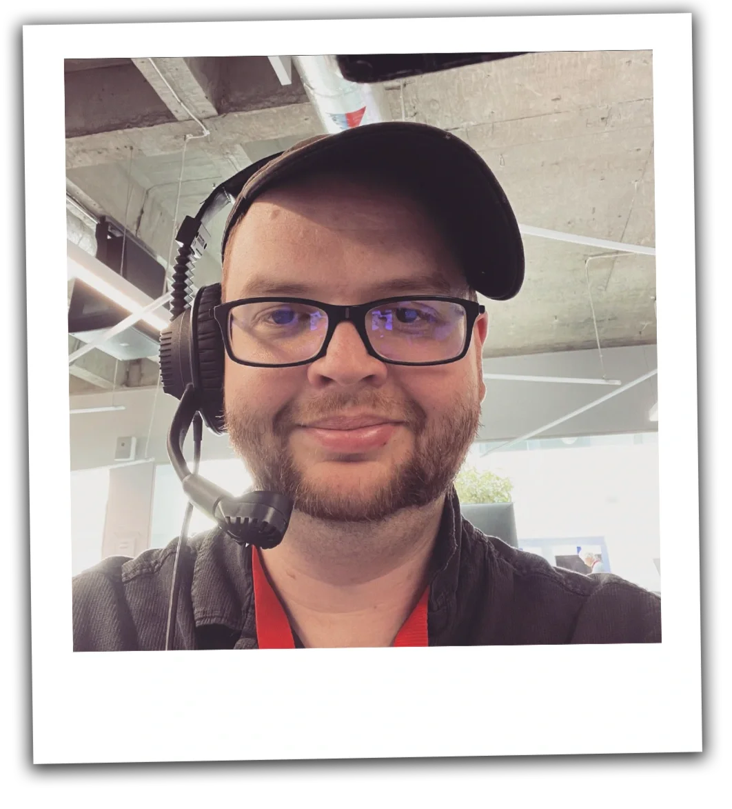 A man wearing a baseball cap, glasses, and a headset, smiling indoors with a modern office background.