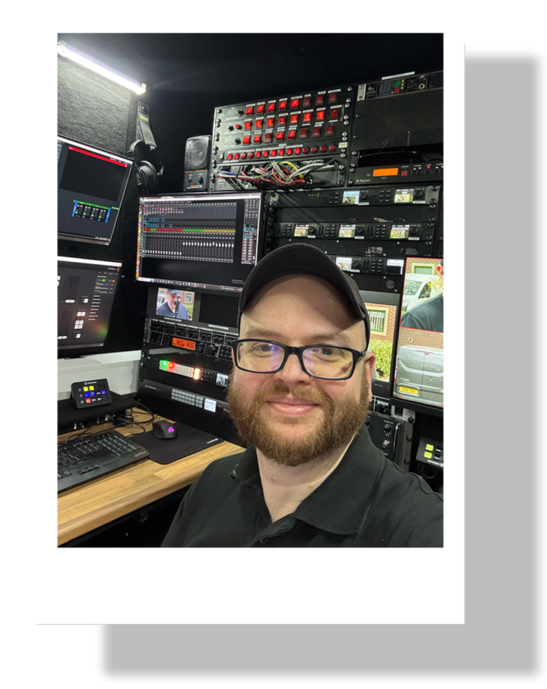 A man with glasses and a cap smiling in a control room filled with audio and video equipment, including monitors, keyboards, and broadcasting gear.