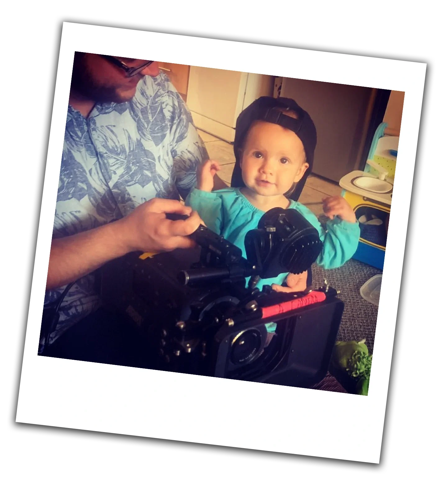 A man filming a young child with a professional camera in a home setting, with the child sitting in a high chair, making a fist, and wearing a baseball cap backwards.