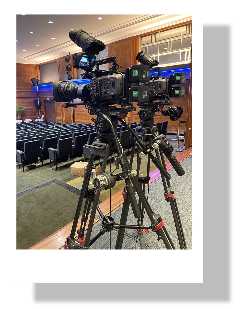 Two professional video cameras on tripods set up in an auditorium with black chairs, wooden walls, and a stage, ready for filming.