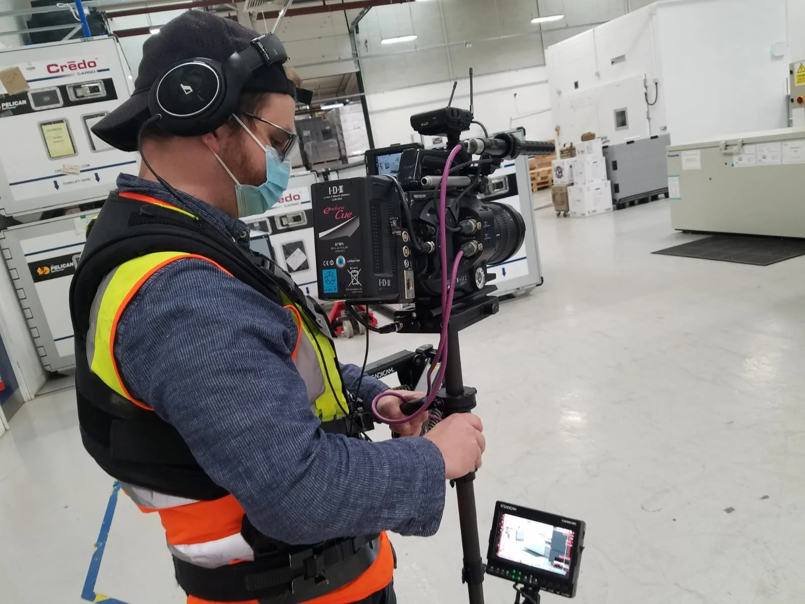 A man wearing a black cap, glasses, a face mask, a blue shirt, and a neon yellow safety vest operates a professional video camera in an industrial warehouse.