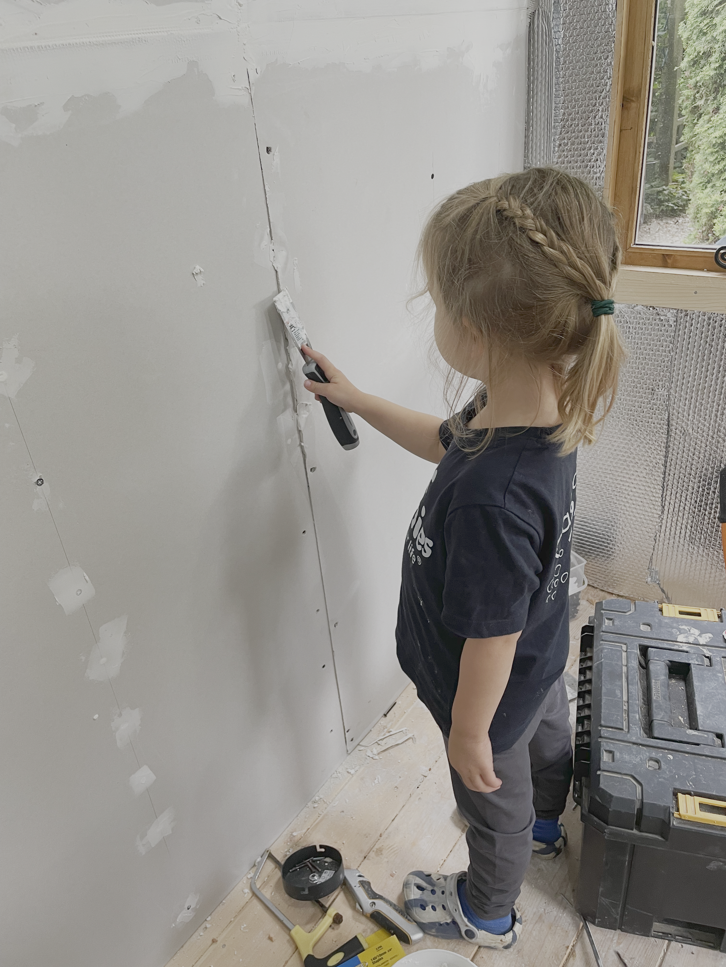 Child using a putty knife to apply joint compound on drywall in a construction setting.