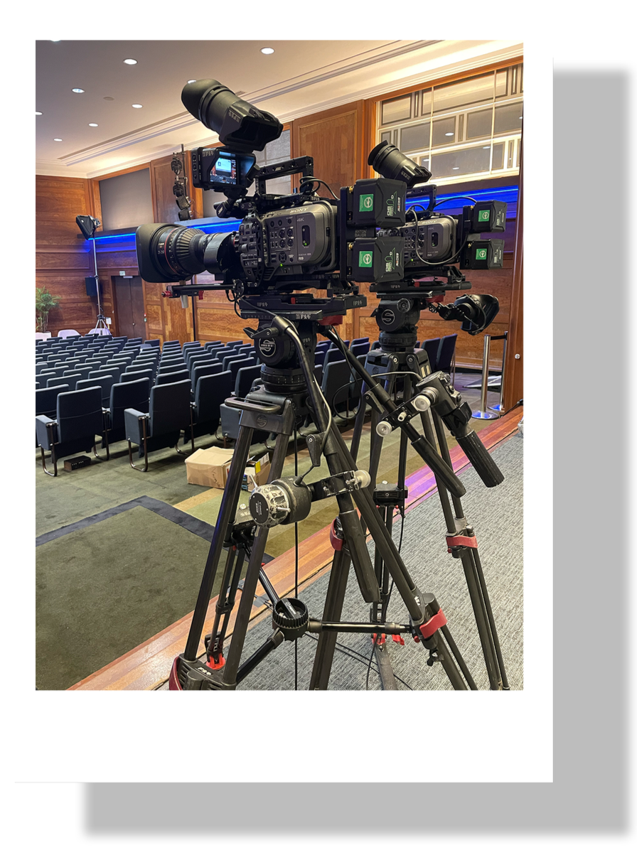 Two professional cameras on tripods set up in an auditorium with rows of empty chairs, wood-paneled walls, and stage lighting.